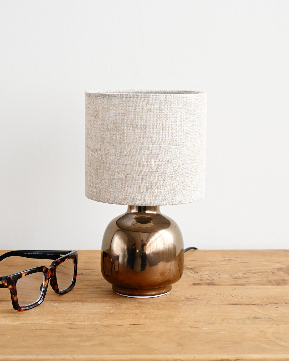 Table lamp with a beige shade on a wooden surface next to a pair of glasses.