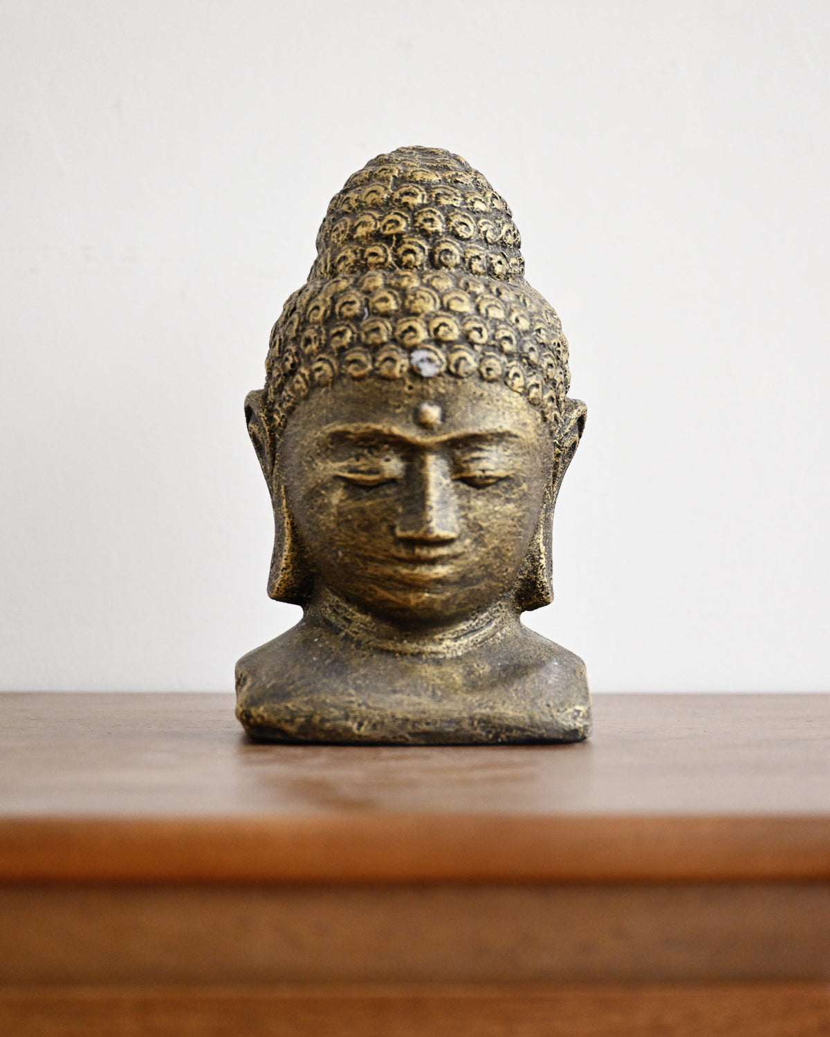 A small bronze Buddha head bust sculpture displayed on a wooden surface against a plain light gray background.