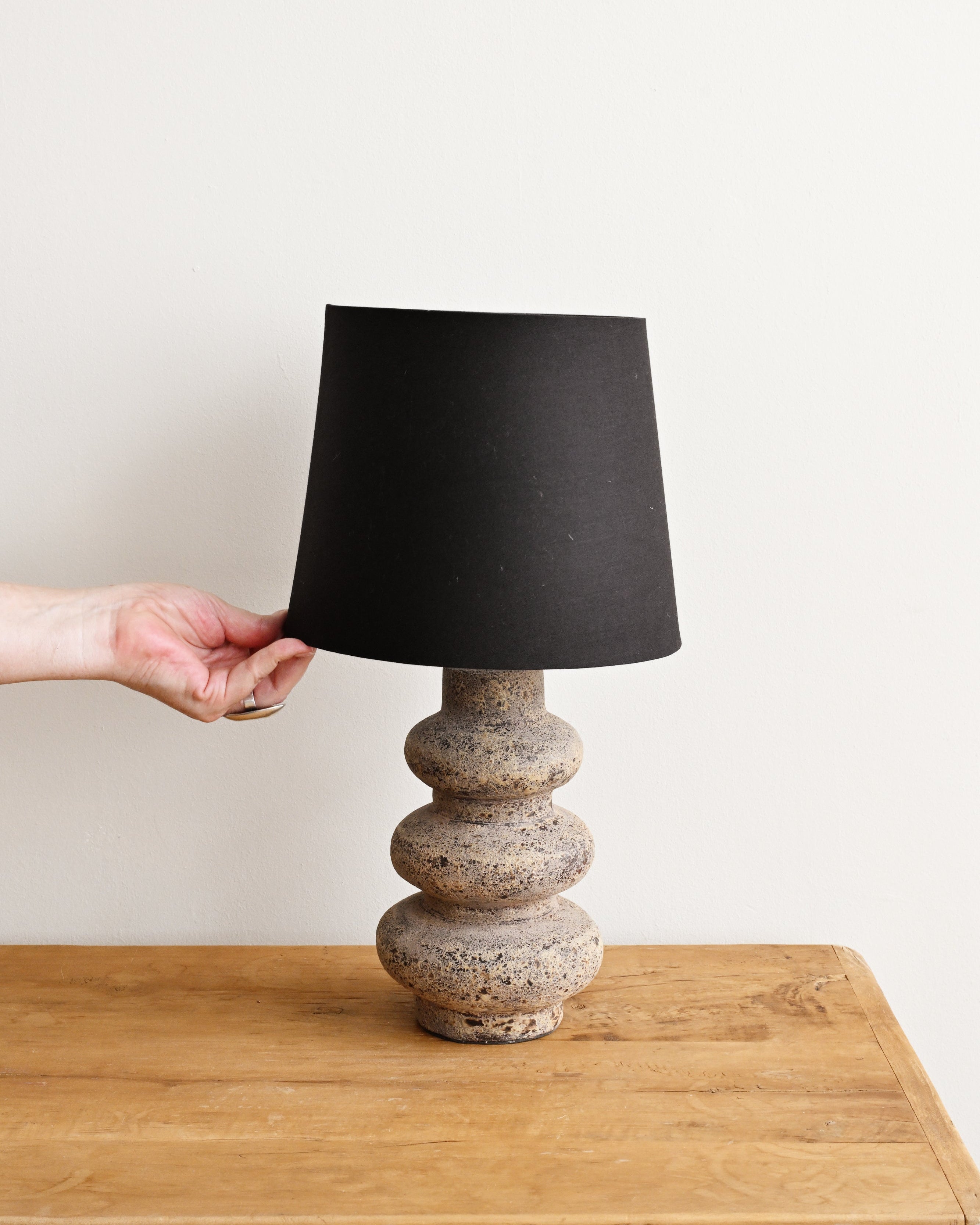 Person holding a black lampshade over a textured ceramic base on a wooden surface.