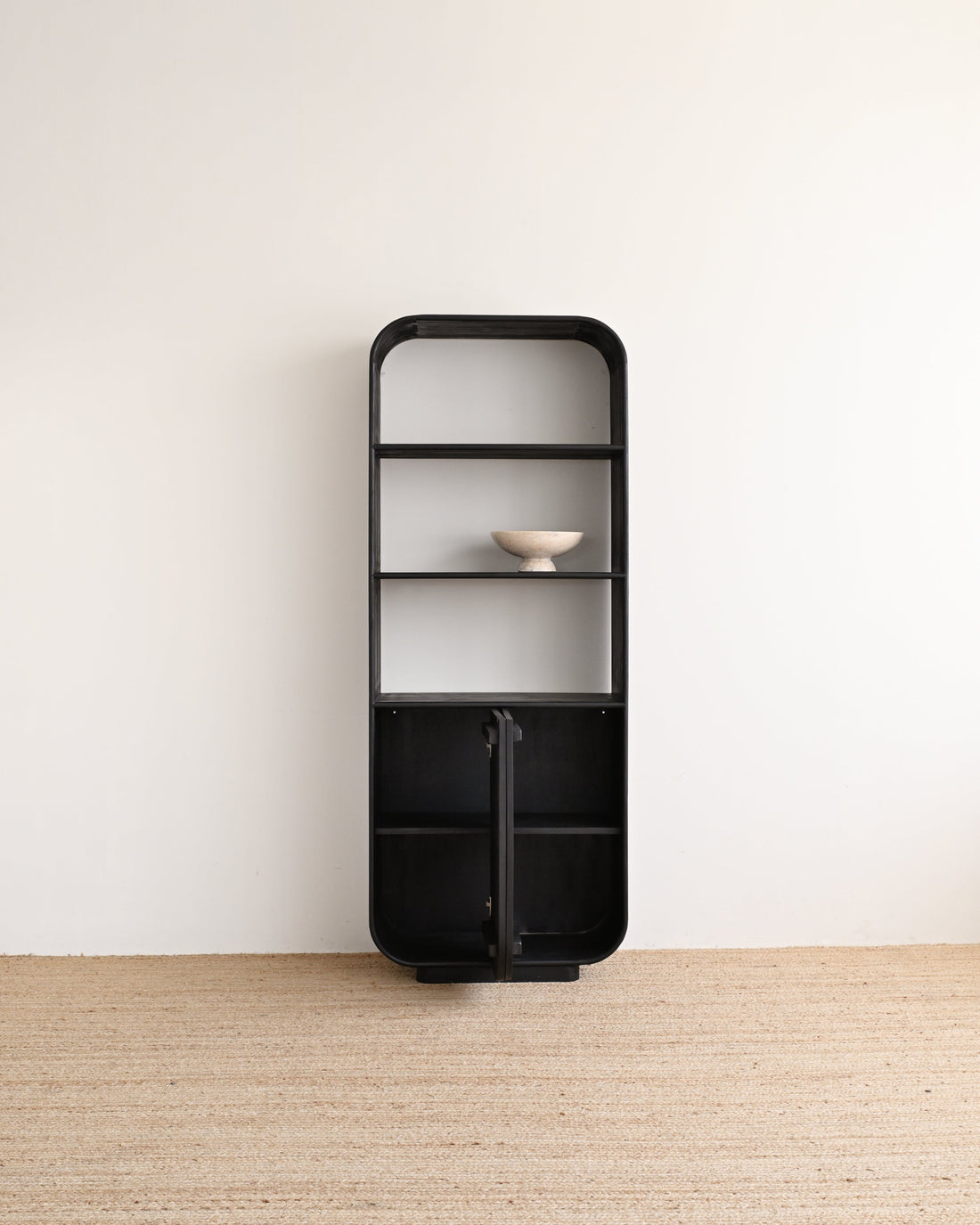 A tall, minimalist black metal shelving unit with a rounded top and bottom, featuring three open shelves and two lower cabinet doors, shown against a plain off-white wall and light beige woven floor.