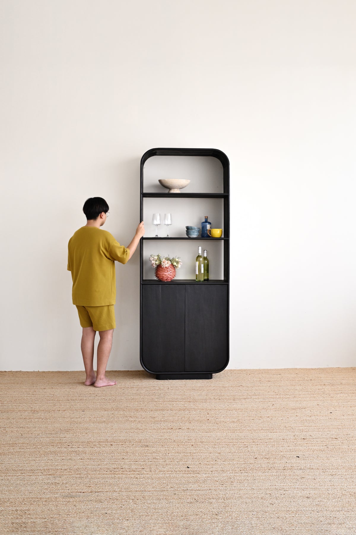 A person in a yellow-green shirt and shorts stands in front of a tall black shelving unit in a minimalist room with a white wall and light beige floor.