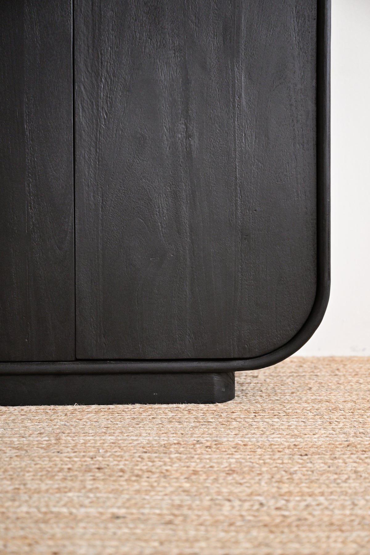 Close-up of a dark charcoal wood cabinet corner with rounded edges and a black metal frame, standing on a textured beige jute rug against a white wall.