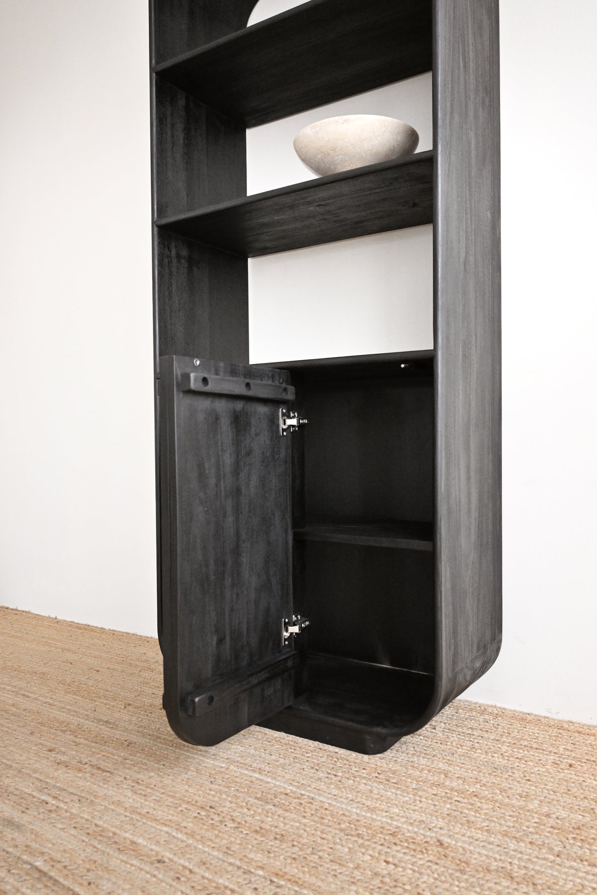A tall black wooden shelving unit with an open lower cabinet door, shown in a minimalist room setting with a light-colored wall and a woven jute rug.