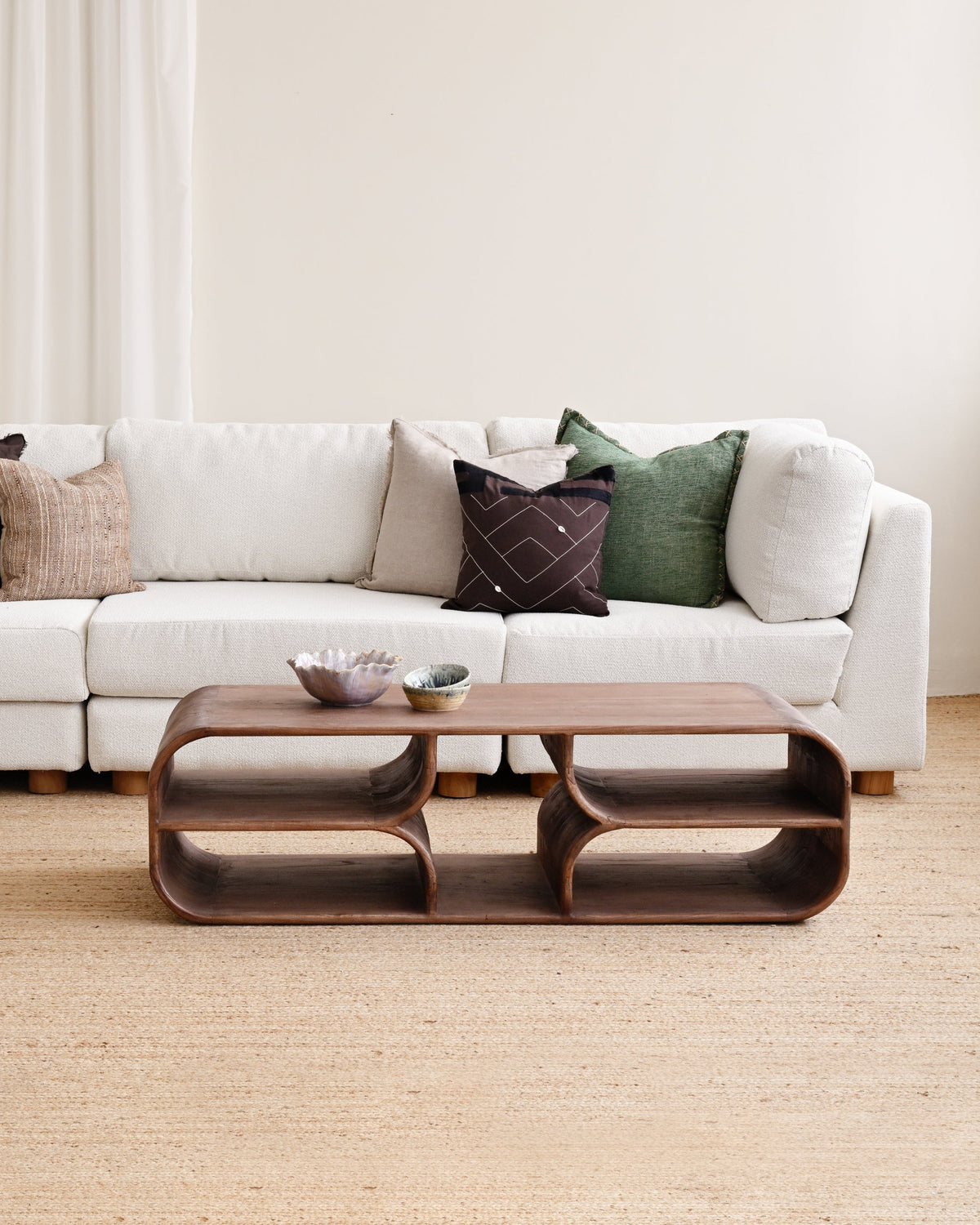 A modern living room setup featuring a white sectional sofa with multiple accent pillows, a matching white ottoman, and a sculptural dark brown wooden coffee table on a light beige jute rug.