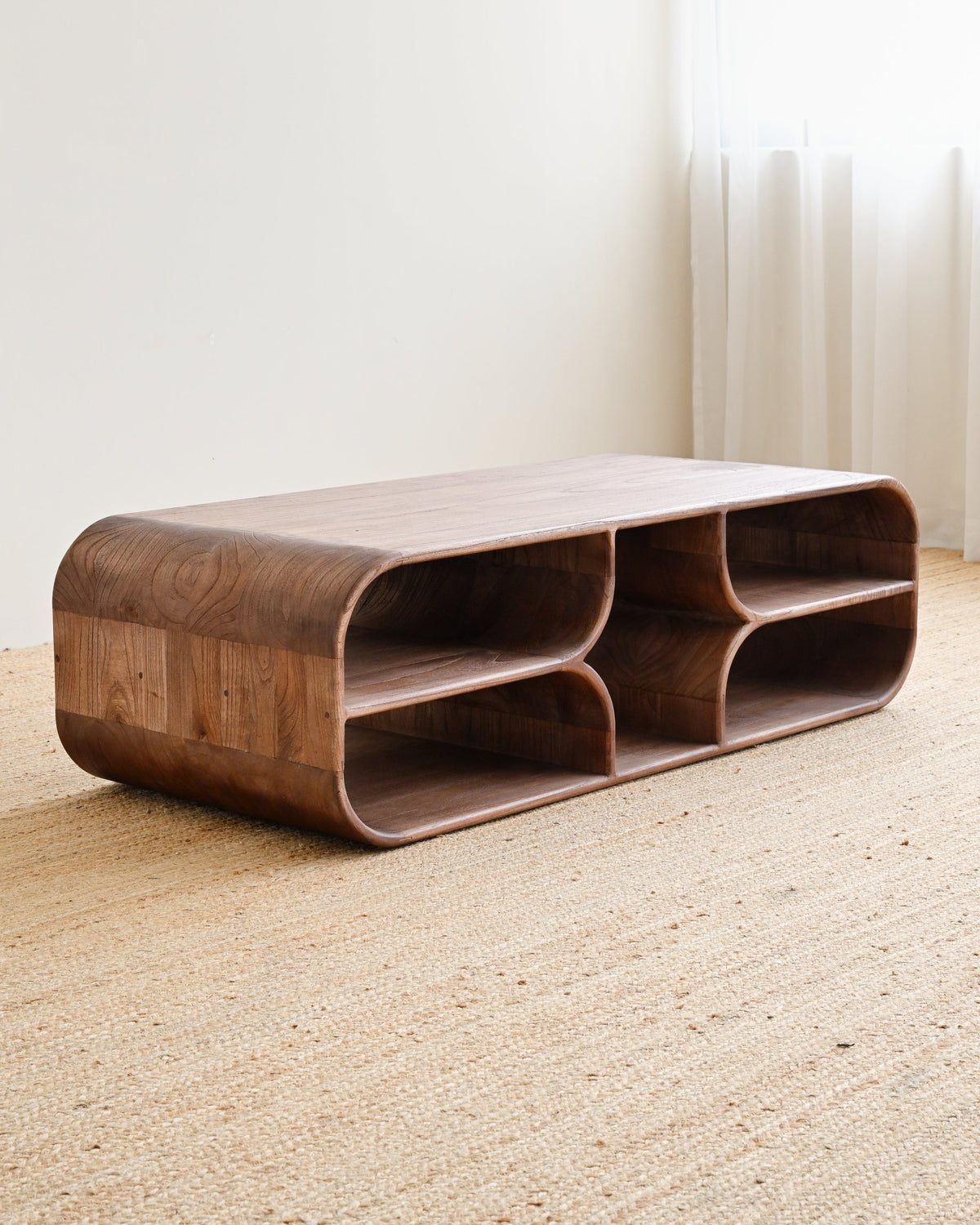 A modern low wooden coffee table with curved, rounded edges and open cubby shelves, shown on a woven jute rug in a minimalist room setting.
