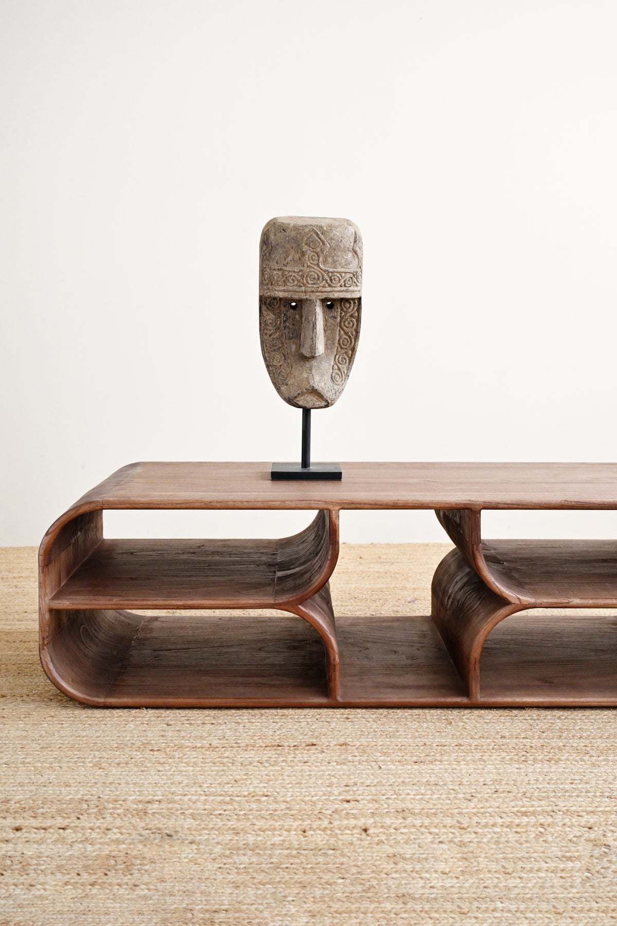 A carved stone mask on a black stand displayed on top of a curved wooden coffee table with open shelves, set on a woven jute rug against a plain off-white wall.