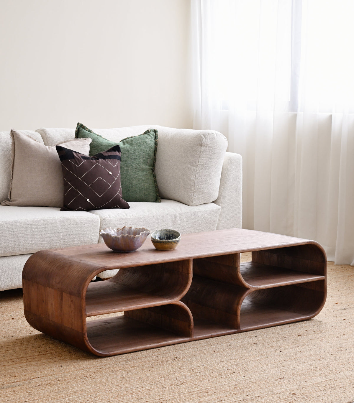 A curved wooden coffee table with open shelves and two small bowls on top, placed in a bright living room in front of a white sofa with decorative pillows.