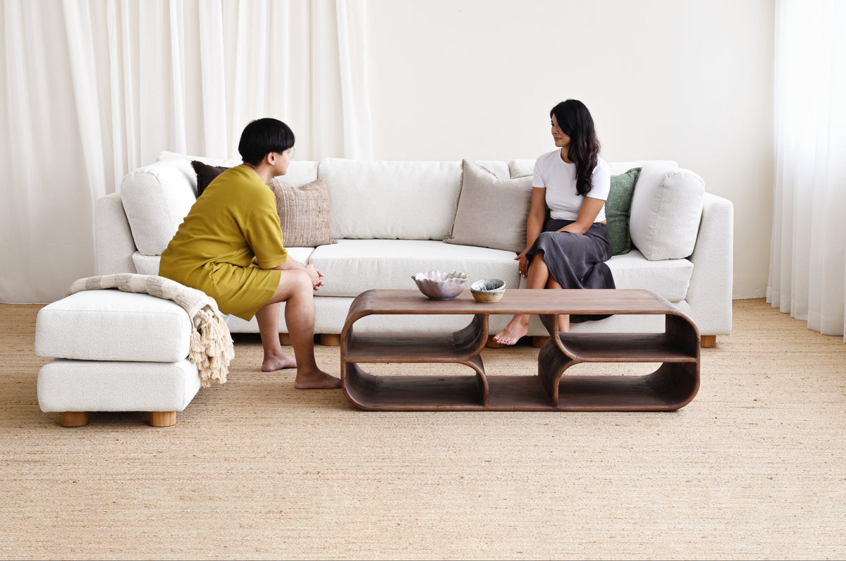 Two women seated on a white sectional sofa in a bright, modern living room, with a dark wood coffee table in front of them and sheer white curtains in the background.