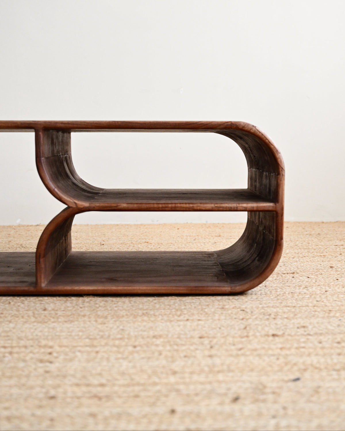 A close-up of a dark brown wooden shelf with a curved, sculptural design, resting on a light beige woven rug against a plain off-white wall.