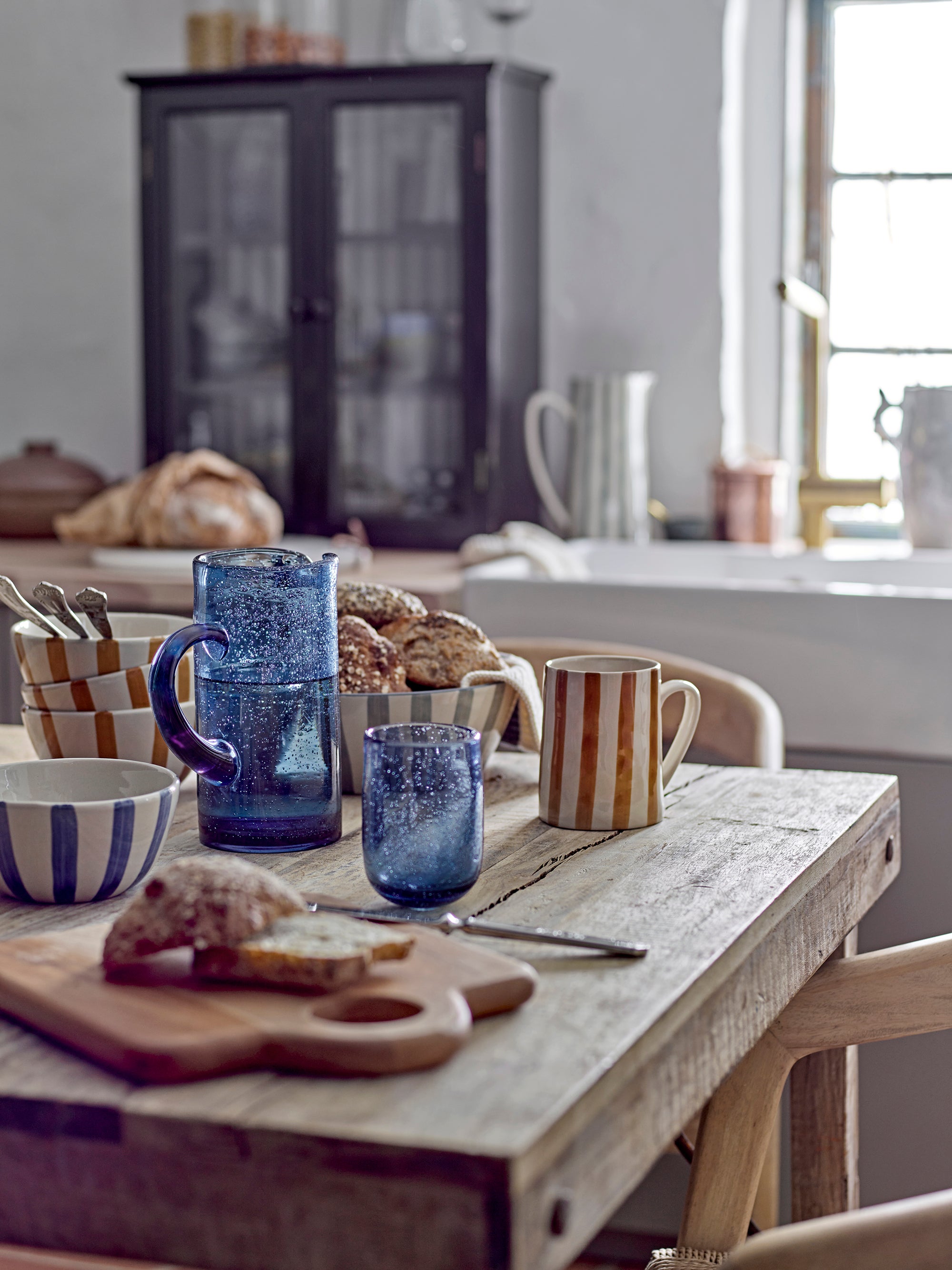 Kitchen table with blue pitcher, striped mug, and bread on a wooden surface.