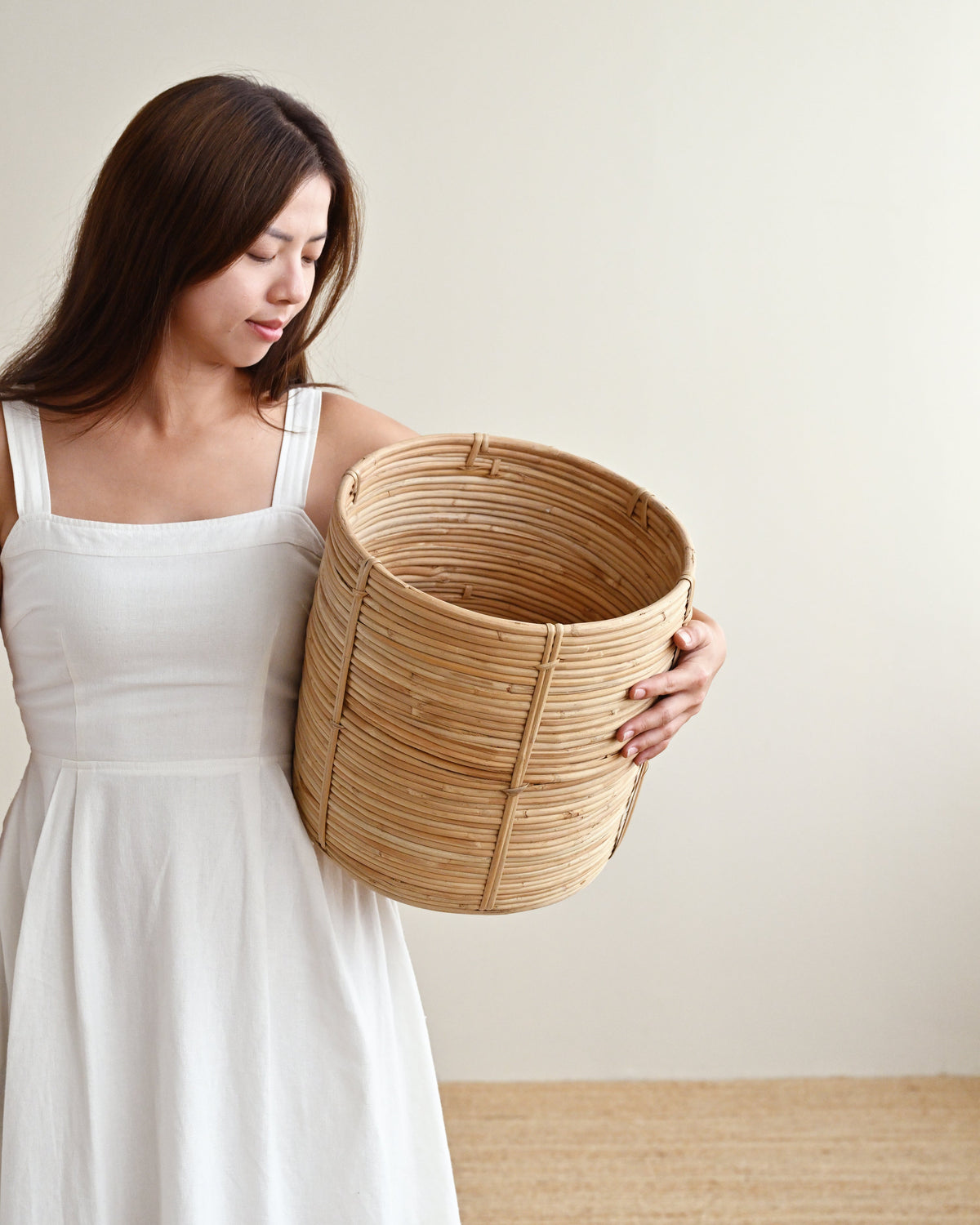 A woman in a white sleeveless dress holds a large round woven basket made of natural light brown wood or rattan, posed against a plain beige wall and light wood floor.