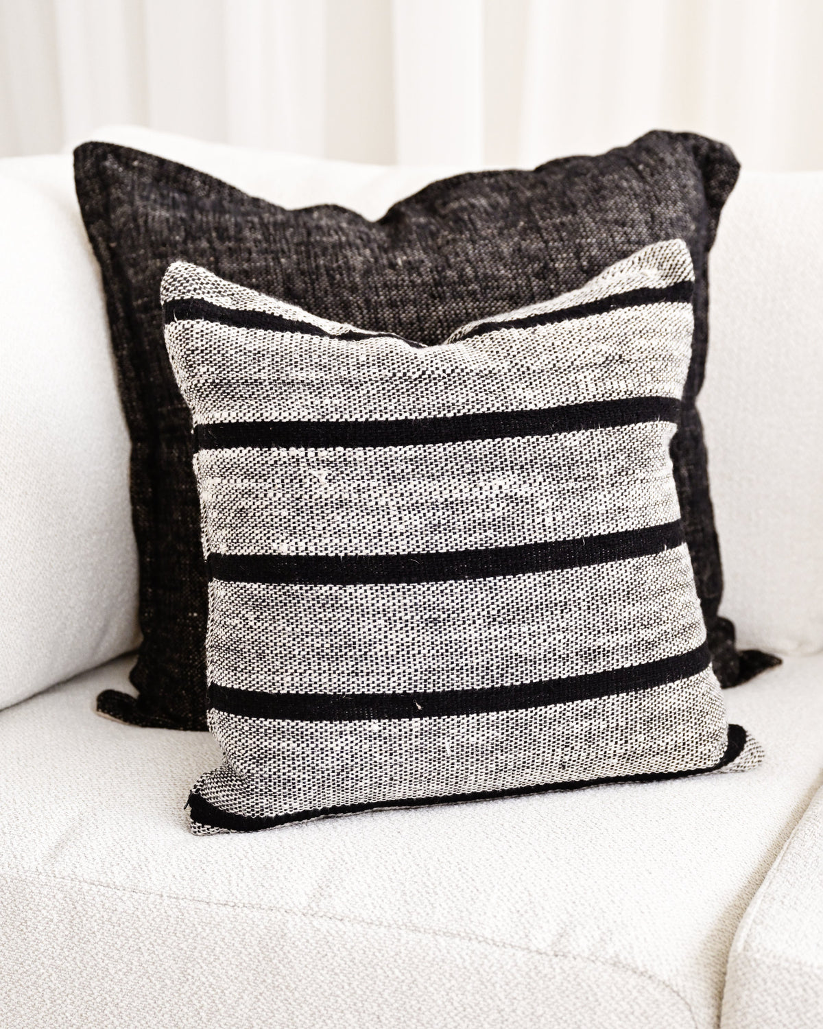 A square throw pillow with horizontal black stripes on a textured gray and white woven fabric, styled on a white sofa in front of a dark charcoal pillow.