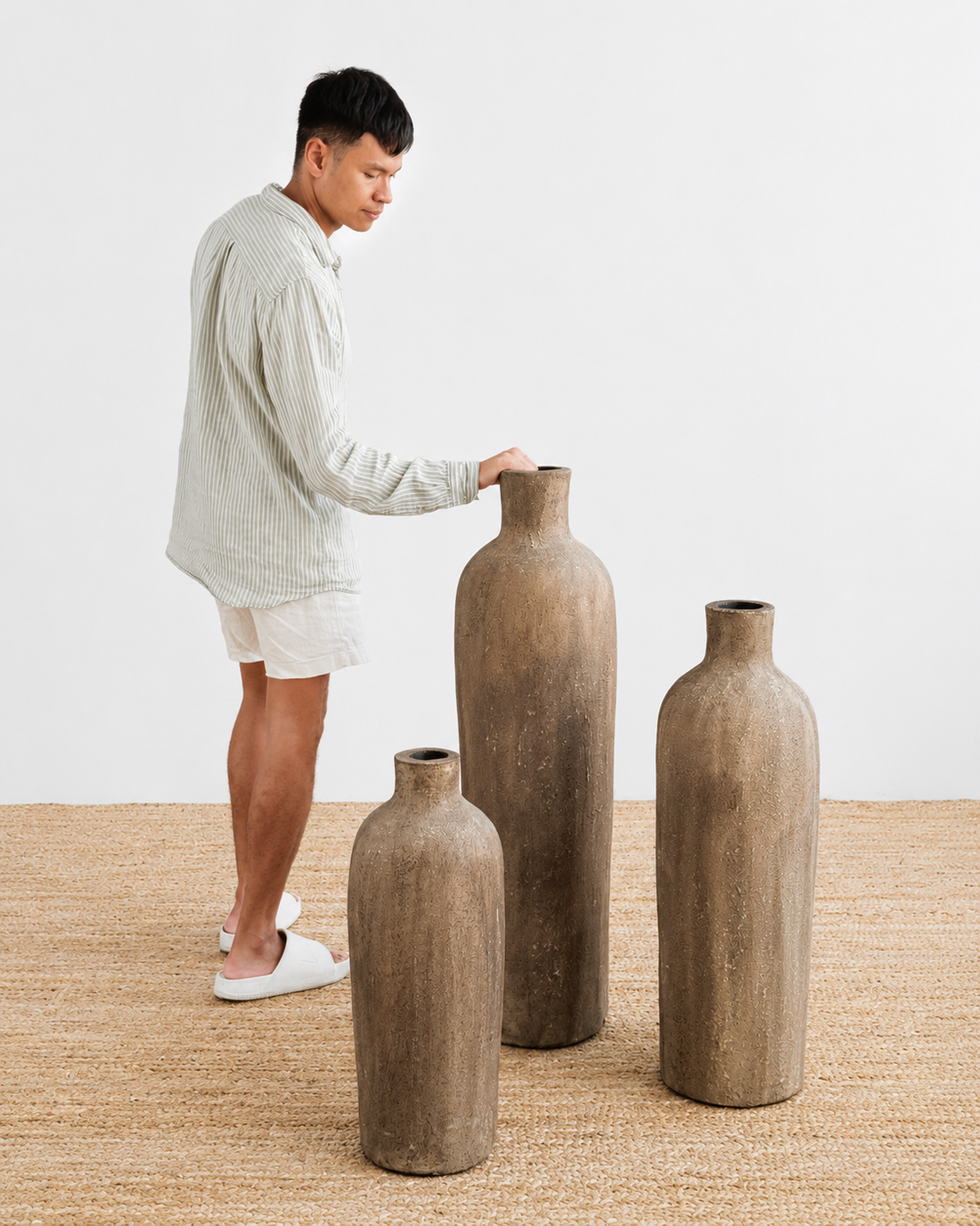 Man arranging large brown vases on a wooden floor with a plain white background