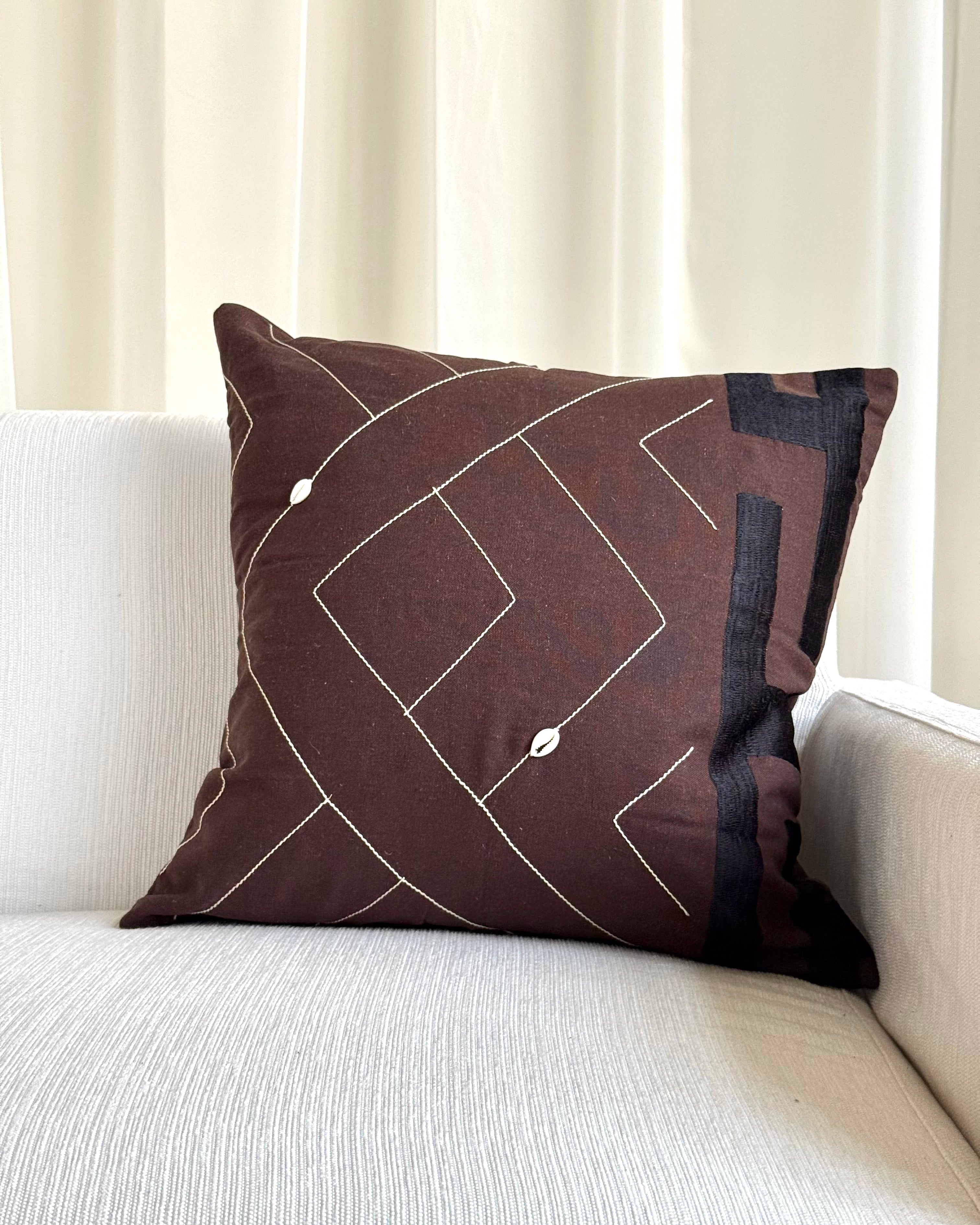A square brown throw pillow with a white geometric line pattern and small white bead accents, resting on a light beige textured sofa in front of cream-colored curtains.