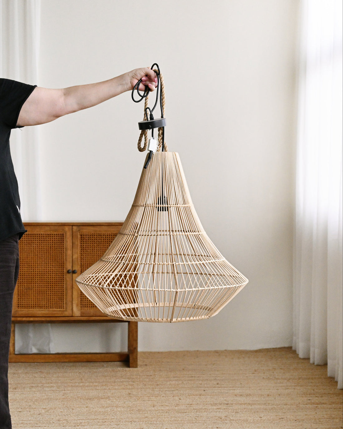 Person holding a wicker pendant light in a room with wooden furniture and white walls.