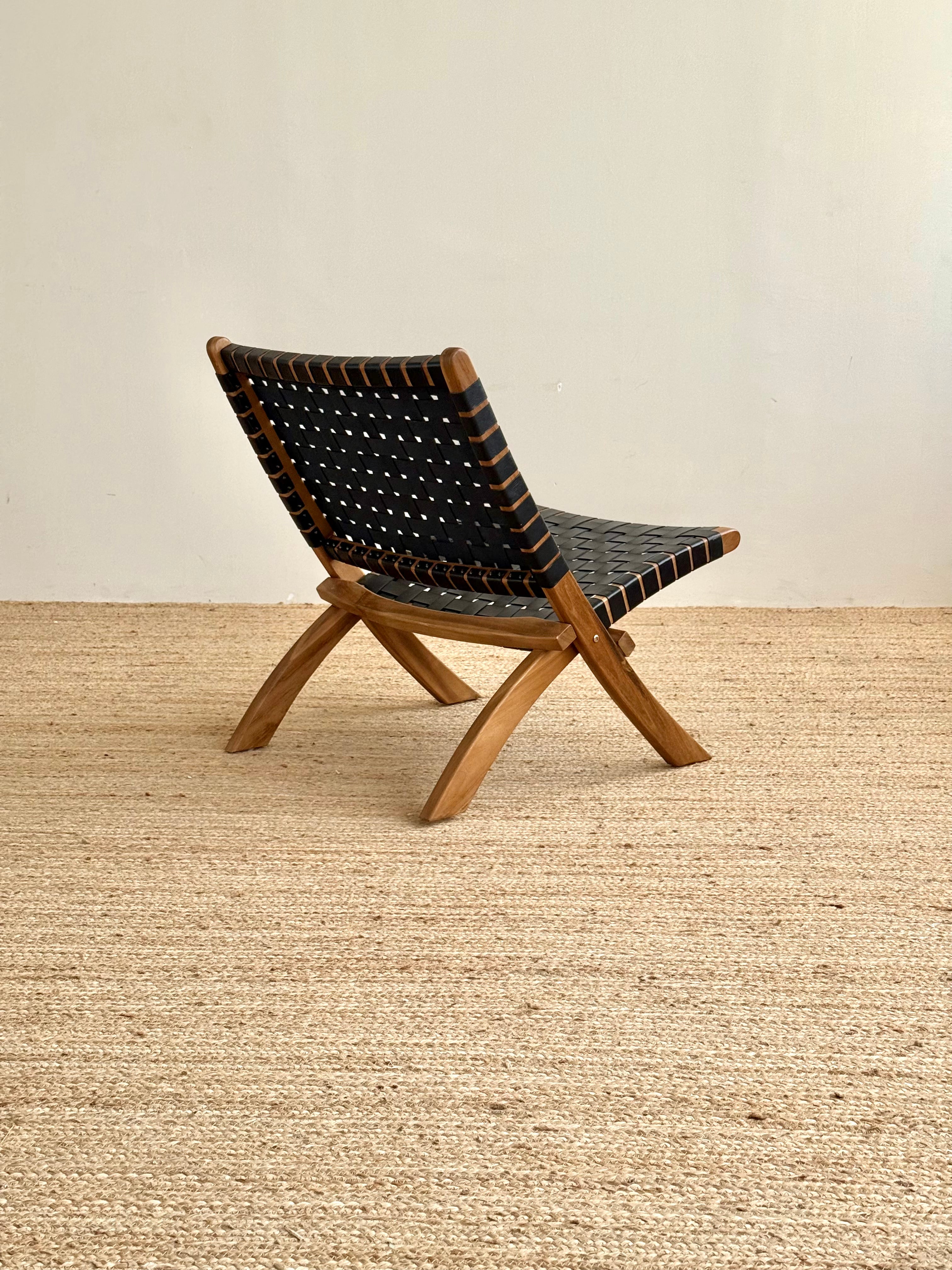 A mid-century modern folding chair with a natural wood frame and black woven leather straps, shown on a textured jute rug against a neutral beige wall.