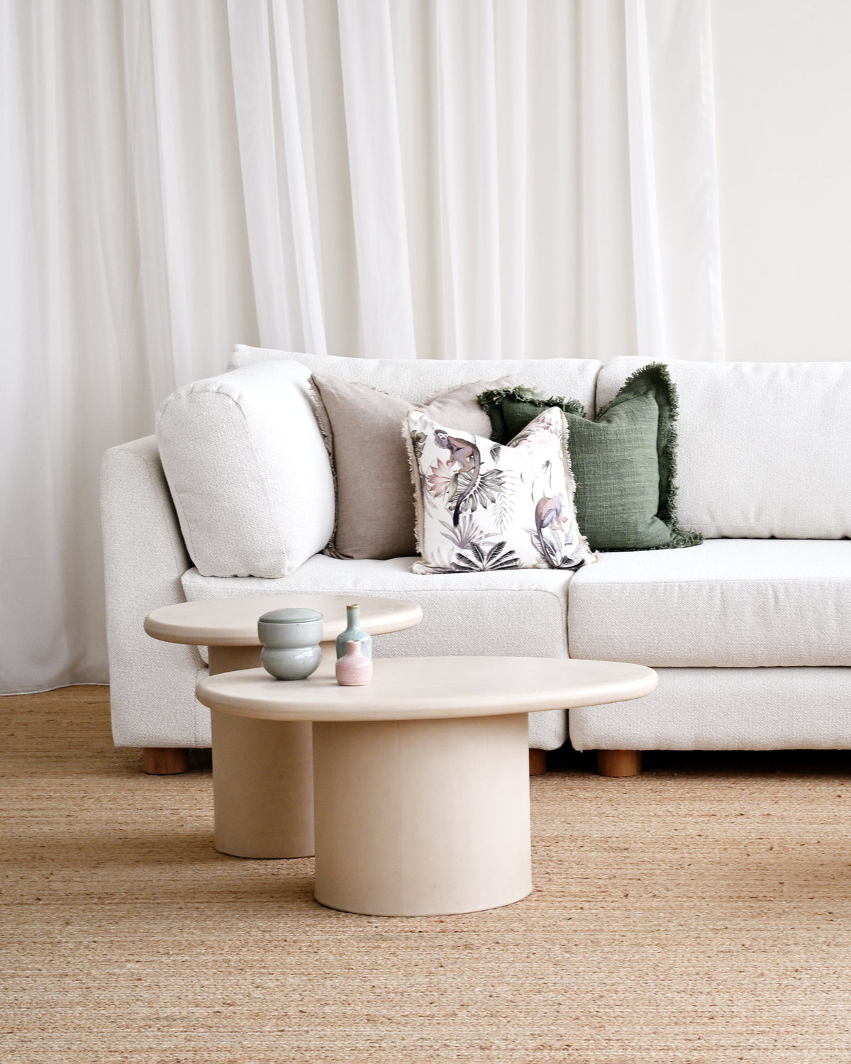 A modern living room setup featuring a white fabric sofa with patterned throw pillows, accompanied by three minimalist round coffee tables in a warm cream/beige tone on a natural jute rug.