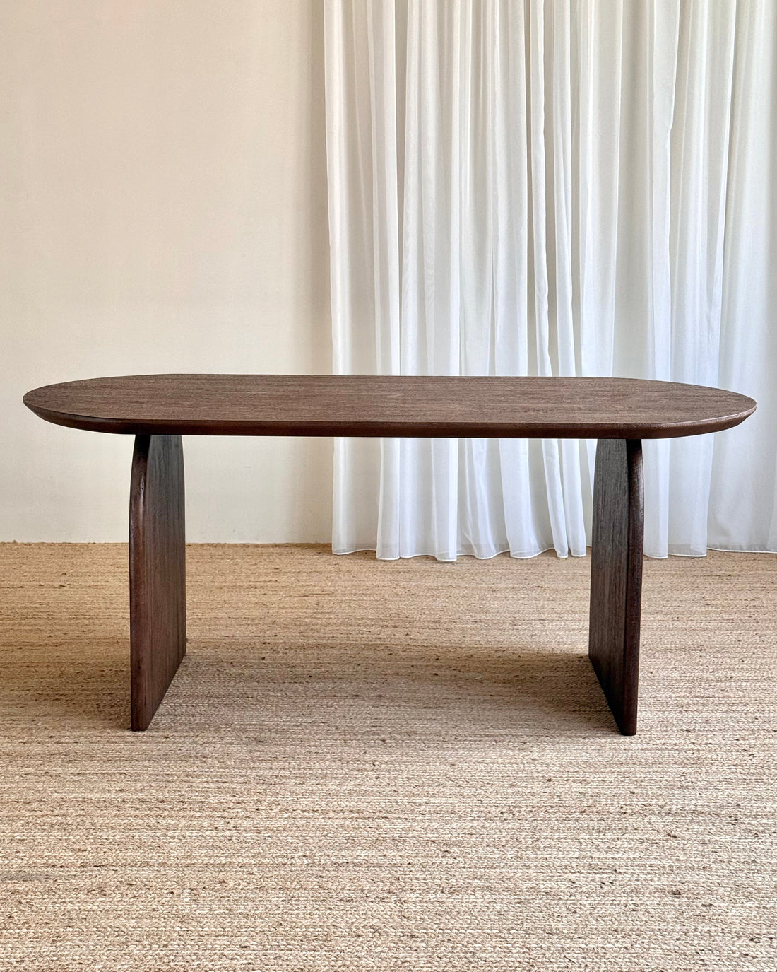 A dark brown wooden dining table with a rounded rectangular top and blocky pedestal legs, set on a textured beige jute rug in front of white curtains.