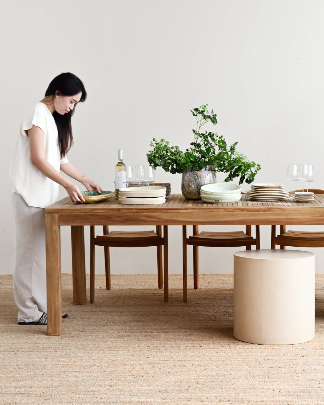 Woman arranging items on a wooden dining table in a minimalistic room with plants.