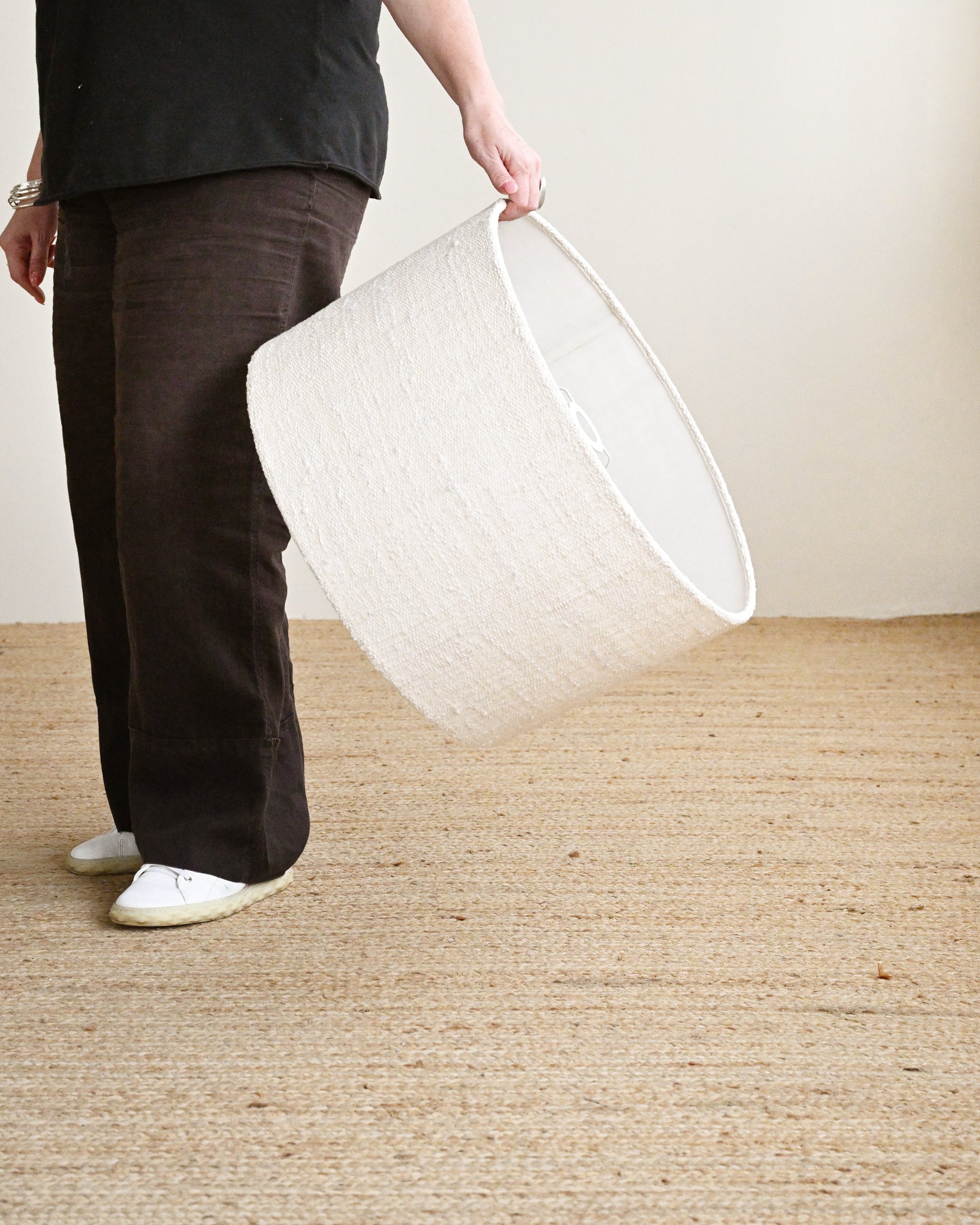 A person holding a white lampshade in a room with a plain wall.