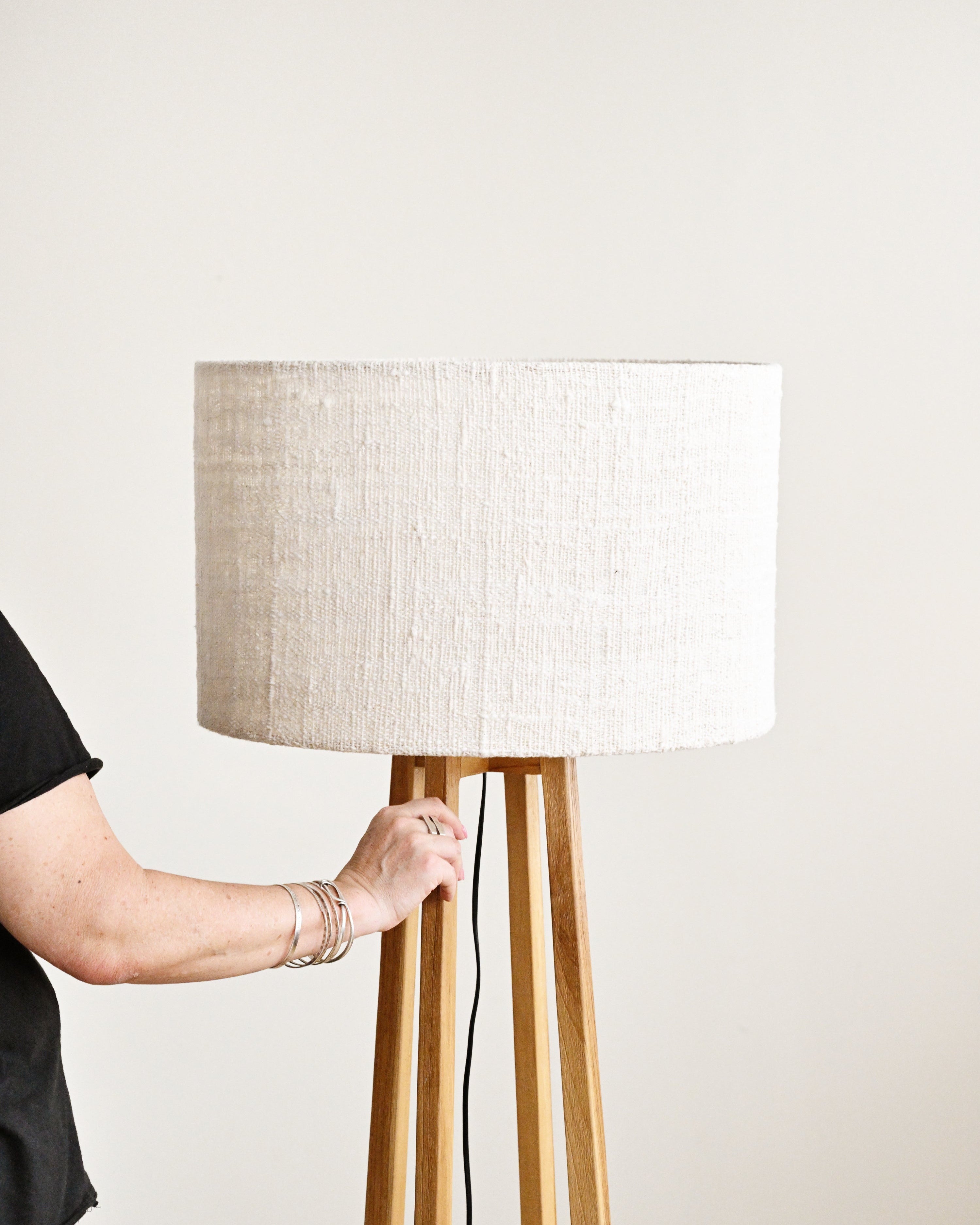 Person adjusting a standing lamp with a white shade and wooden base against a plain background