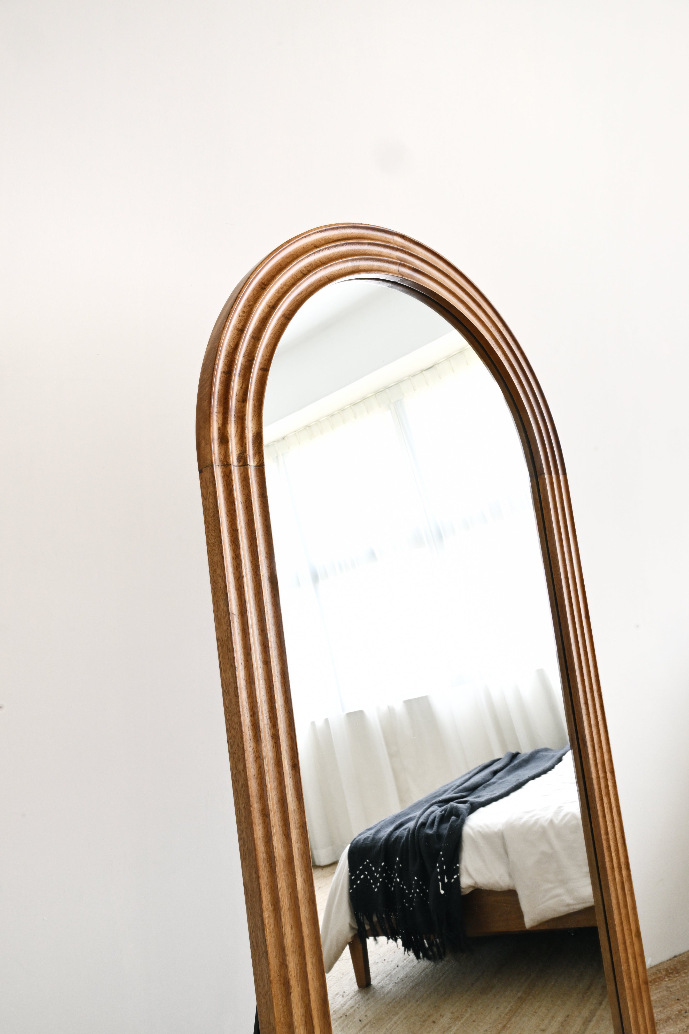 Wooden-framed mirror leaning against a white wall with a bed in the background.