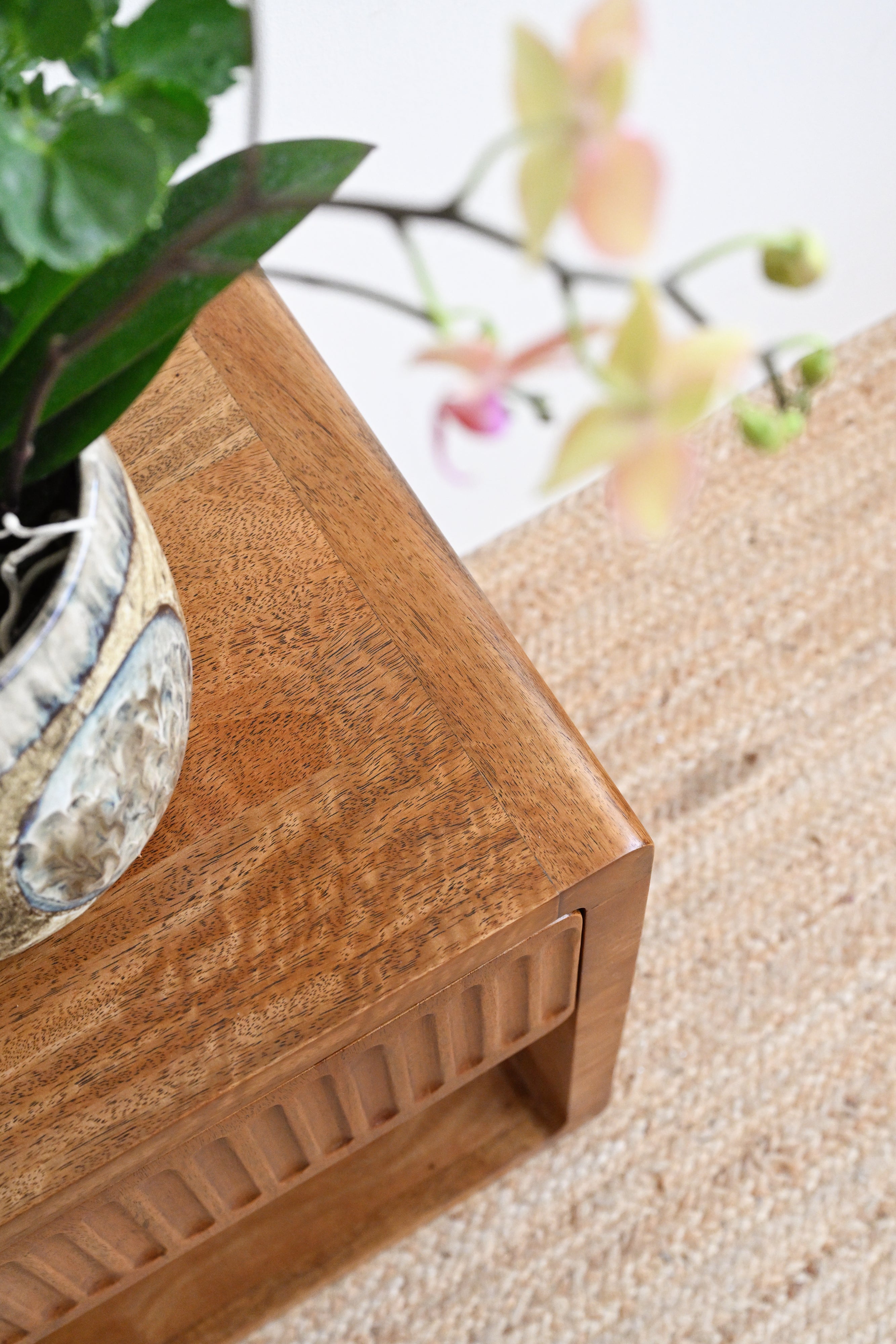 Wooden planter with a plant on a sandy surface with blurred flowers in the background