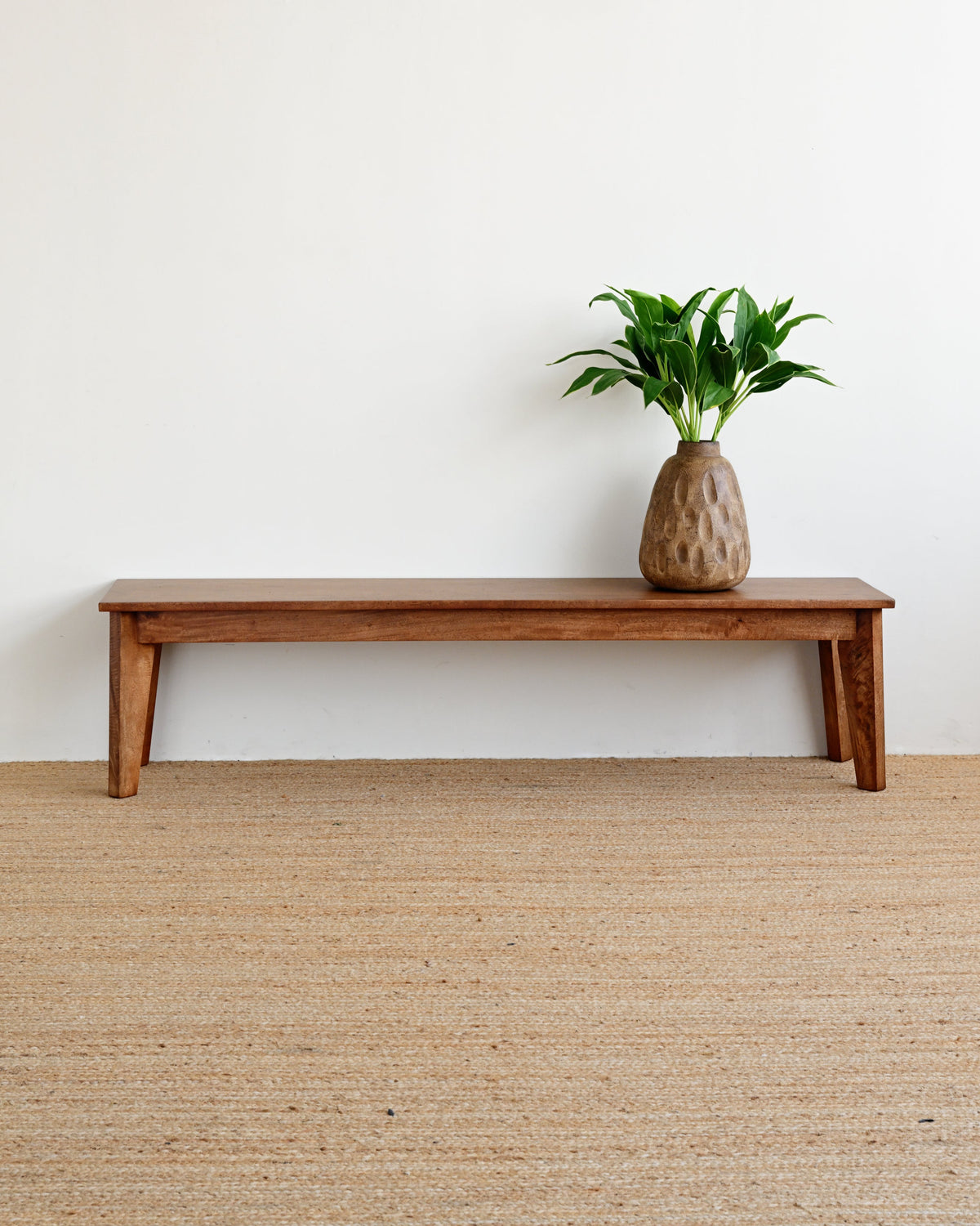 A minimalist wooden bench with a textured ceramic vase containing green leaves, set against a plain white wall and a woven beige rug.
