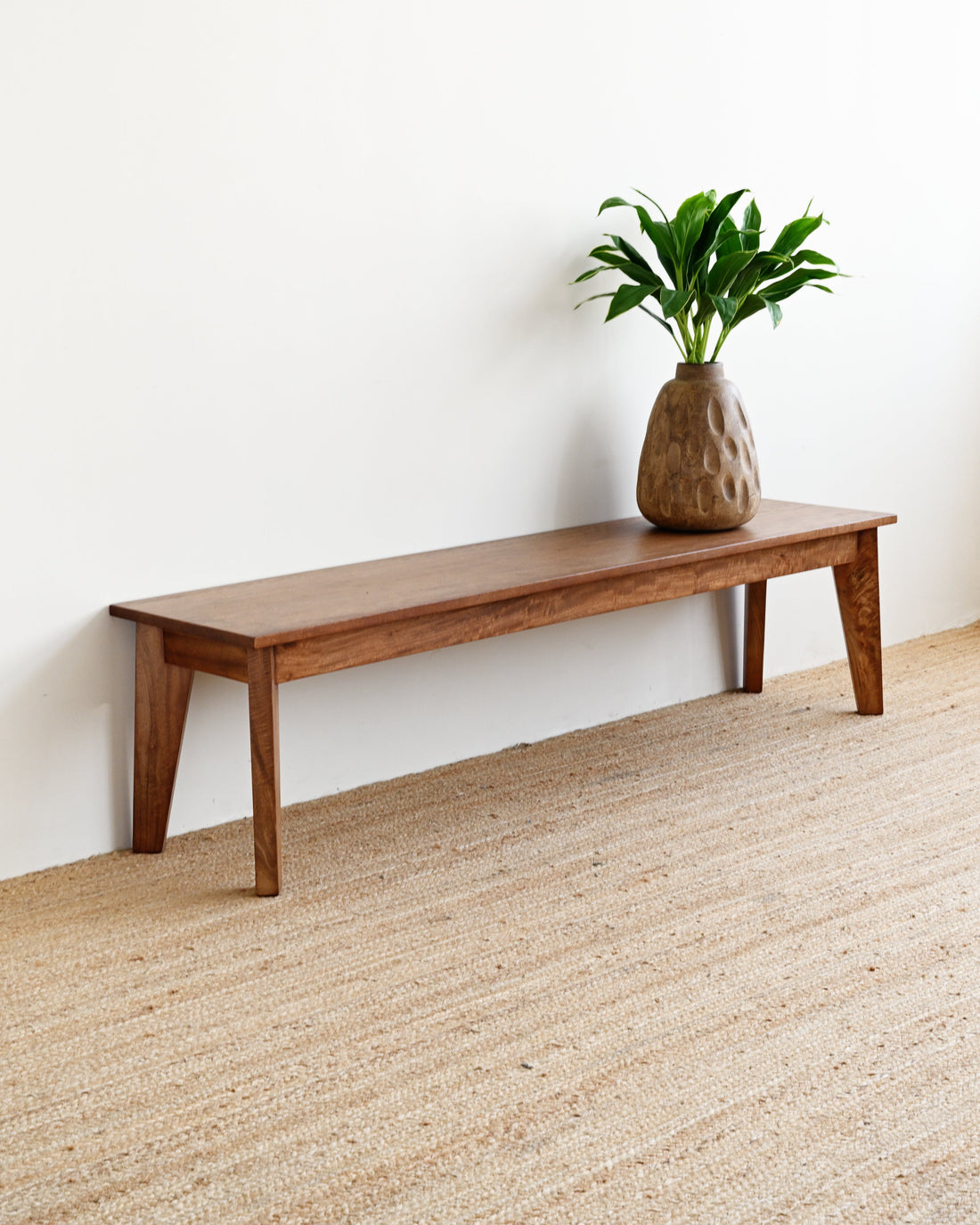 A long wooden bench with angled legs sits against a white wall on a woven jute rug, with a textured brown vase holding a green plant on top.