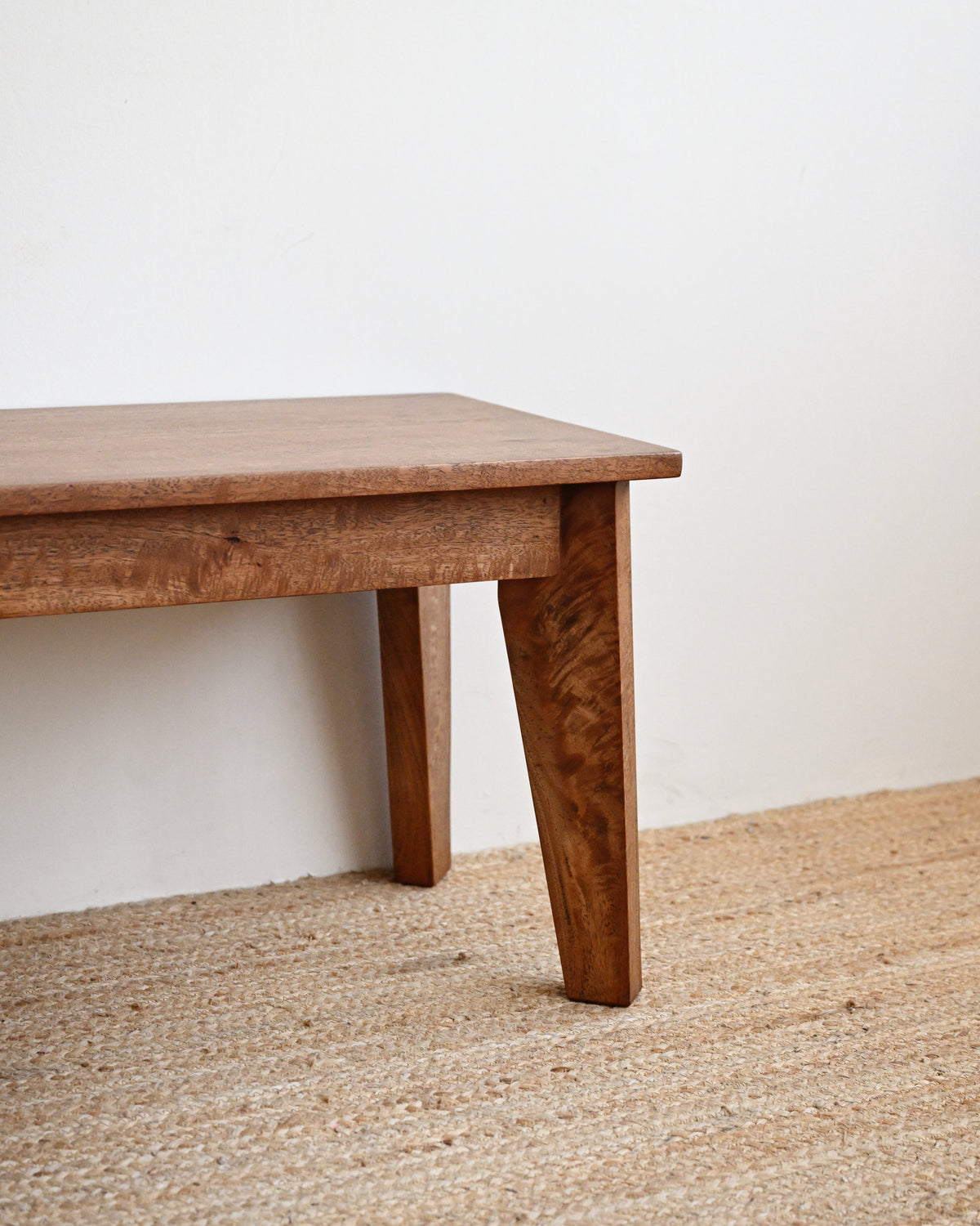 A close-up of a wooden table leg and tabletop corner, showing a medium brown wood grain finish, set against a plain white wall and a natural beige woven jute rug.