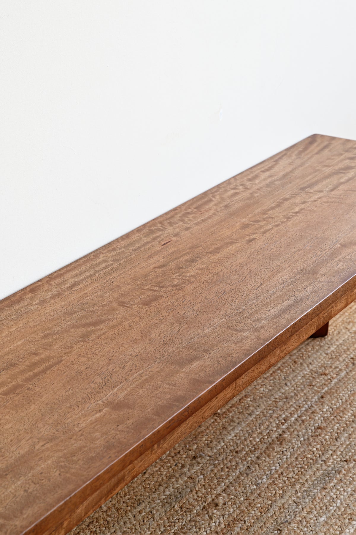 A close-up of a rectangular wooden table or bench with a warm medium-brown wood grain finish, shown resting on a woven jute rug against a plain white wall.