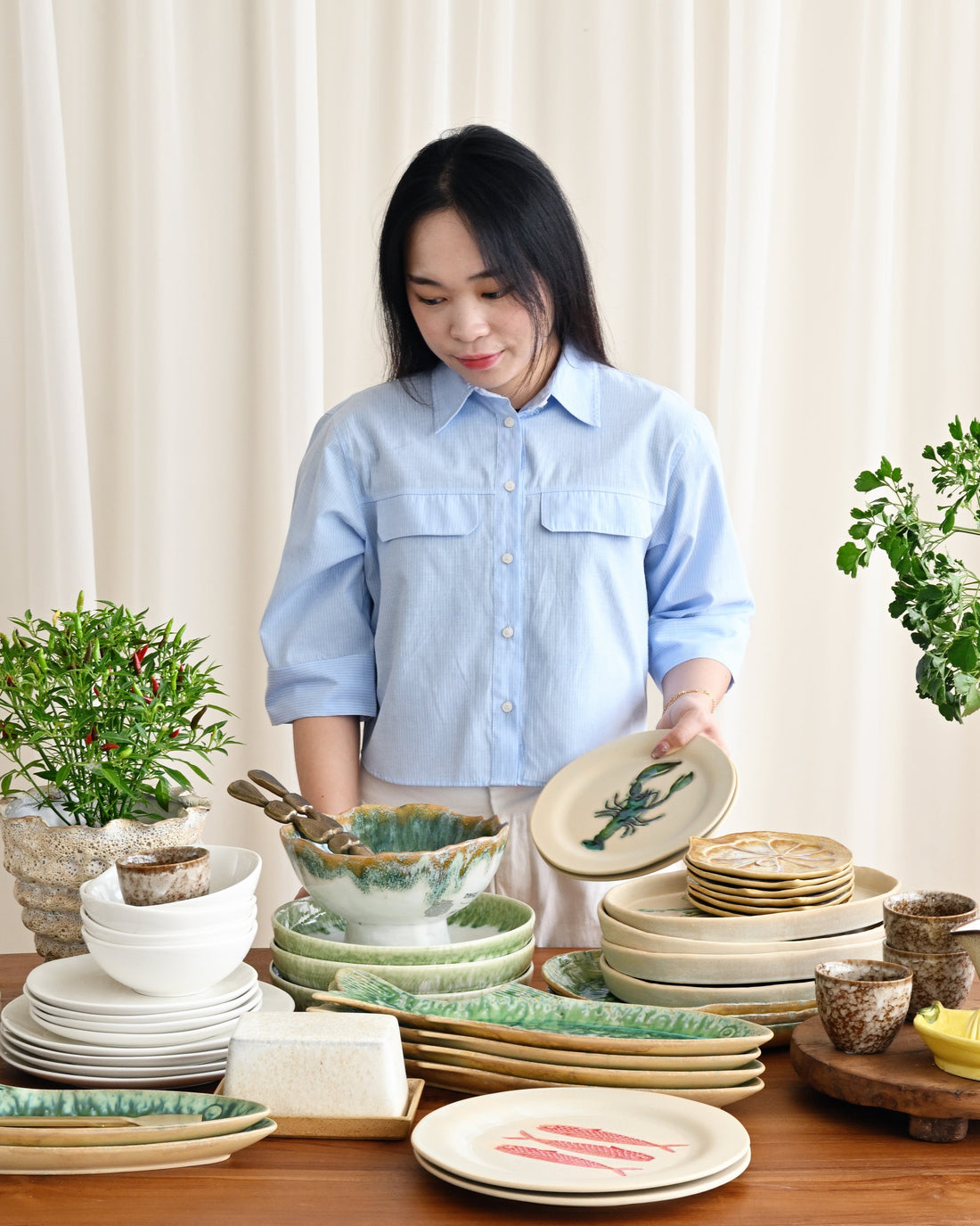 Woman arranging ceramic plates and bowls on a wooden table with plants in the background.