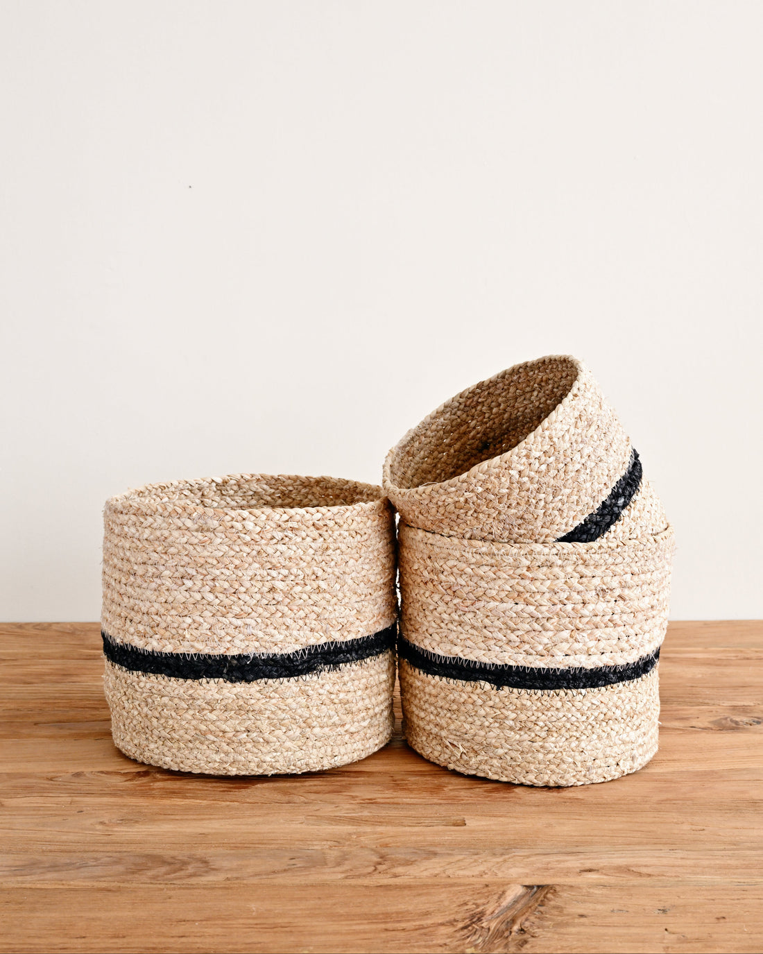 Two woven natural fiber plant baskets with black stripes, one standing upright and one leaning on top, placed on a wooden surface against a plain off-white wall.