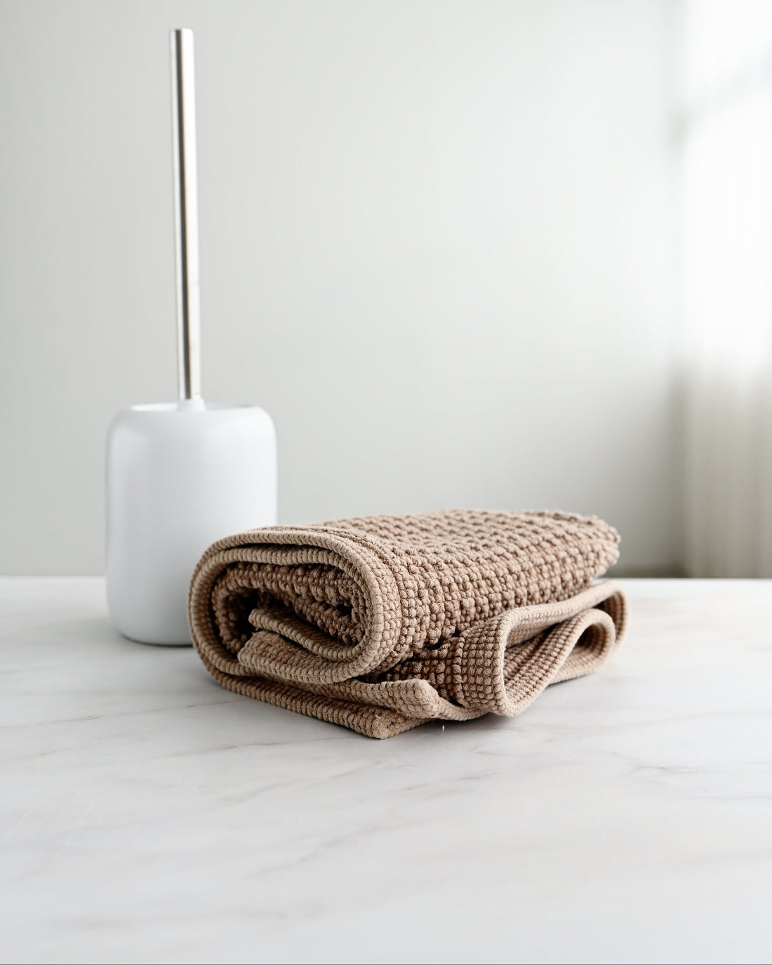 A folded beige waffle-weave throw blanket sits on a light marble surface next to a white ceramic vase with a thin metallic stem, against a soft white background.