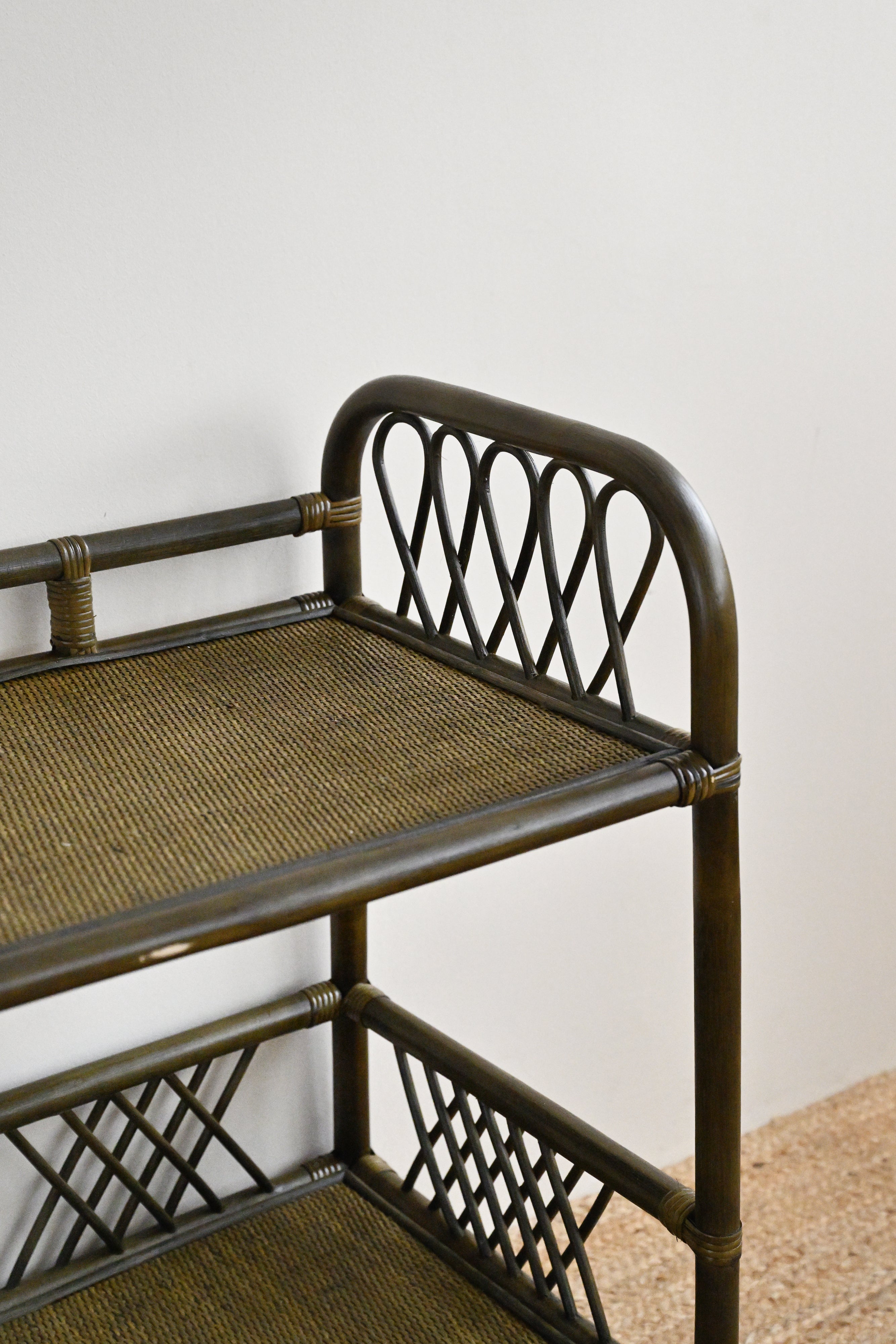 Rattan bar cart with woven seat and shelves against a white wall.