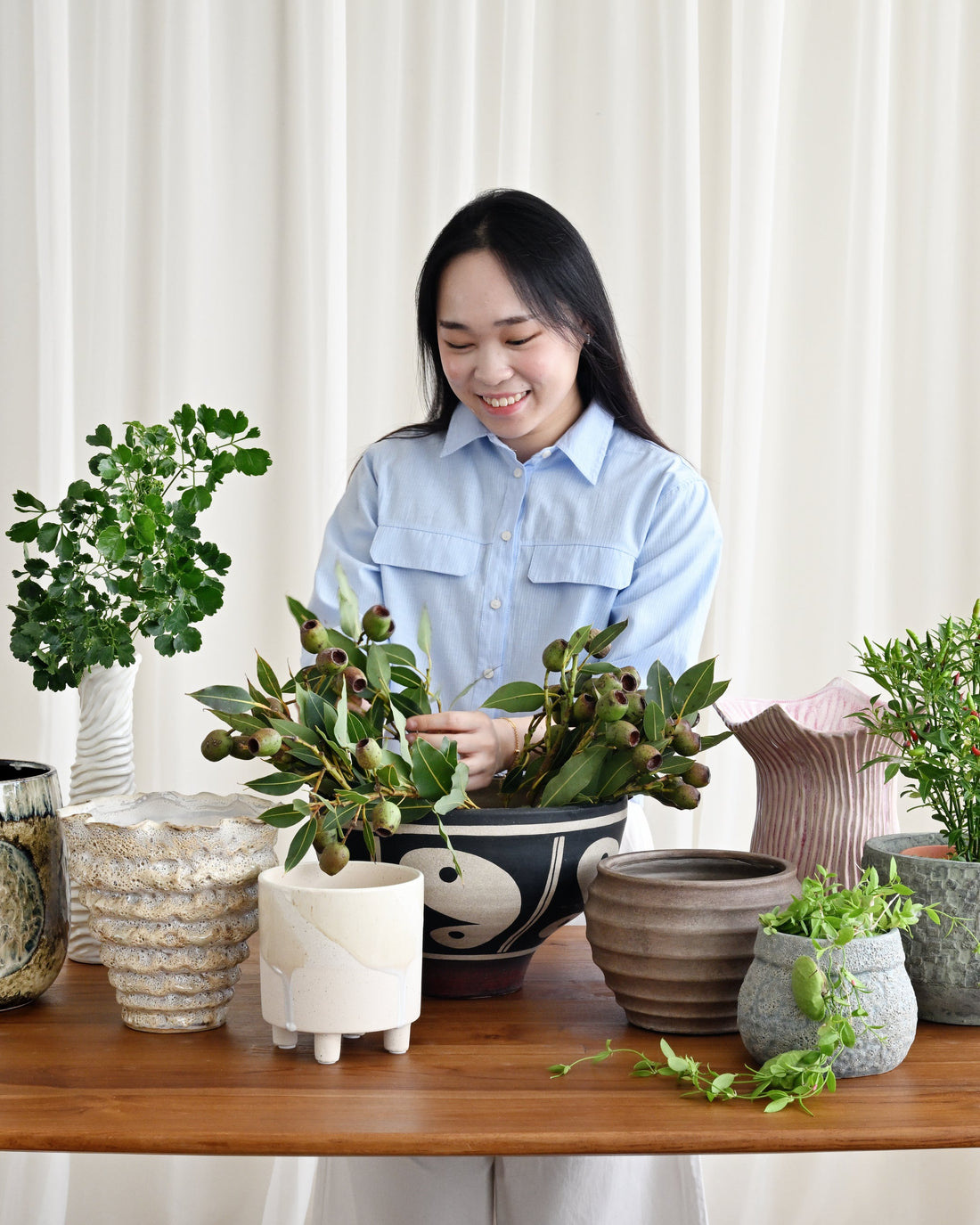 Woman arranging potted plants on a wooden table with a white curtain background