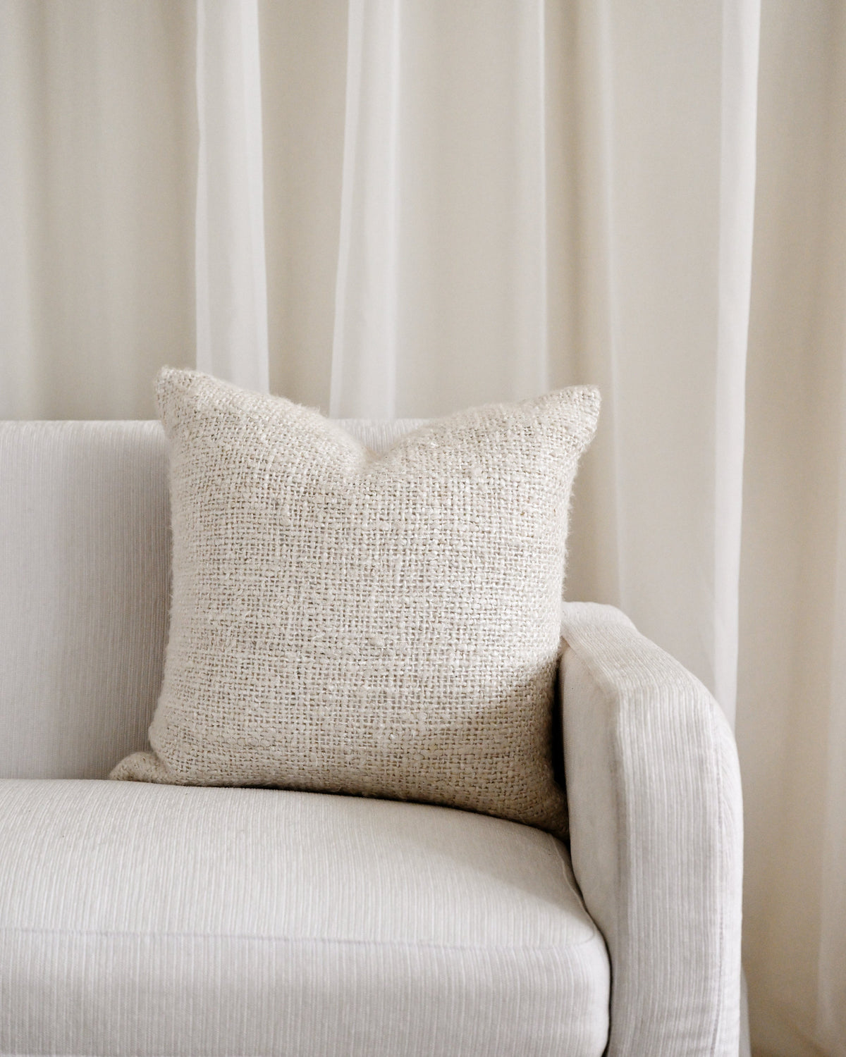 A square beige textured throw pillow resting on a light beige upholstered sofa in front of sheer cream curtains.