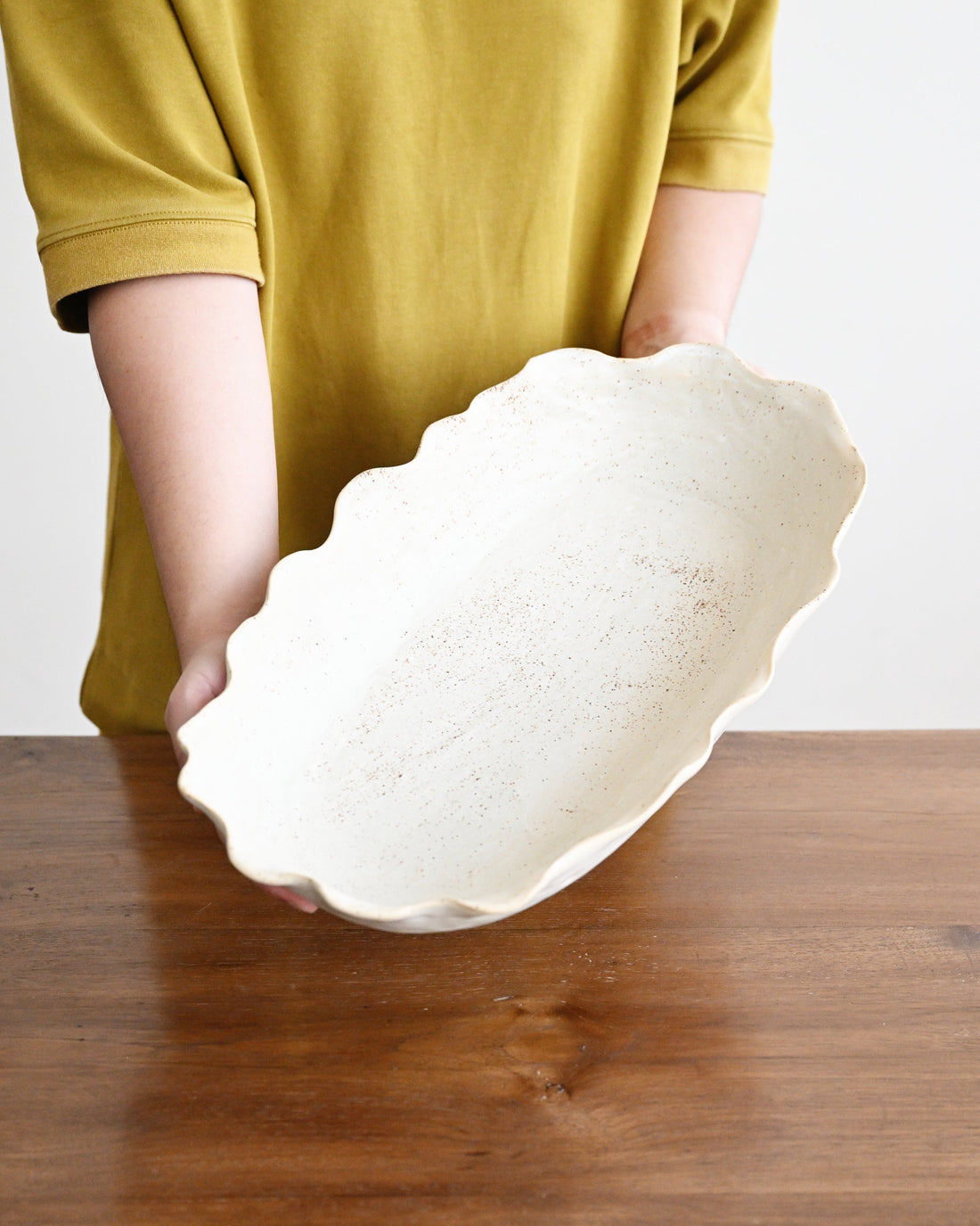 A person wearing a mustard yellow shirt holds a large, scalloped-edge ceramic bowl with a speckled off-white glaze over a dark wooden table.