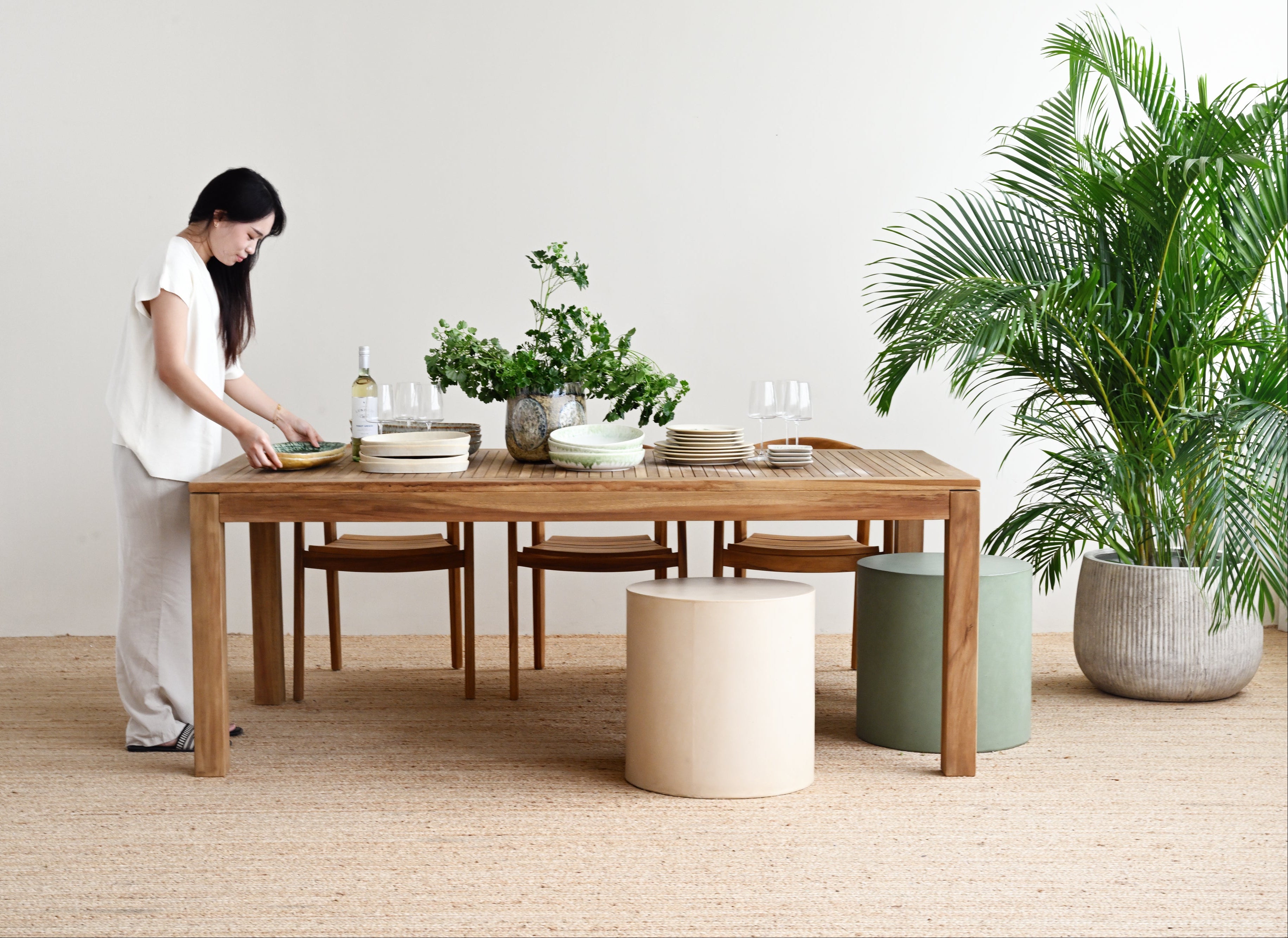 Woman arranging items on a wooden dining table in a minimalistic room with plants.