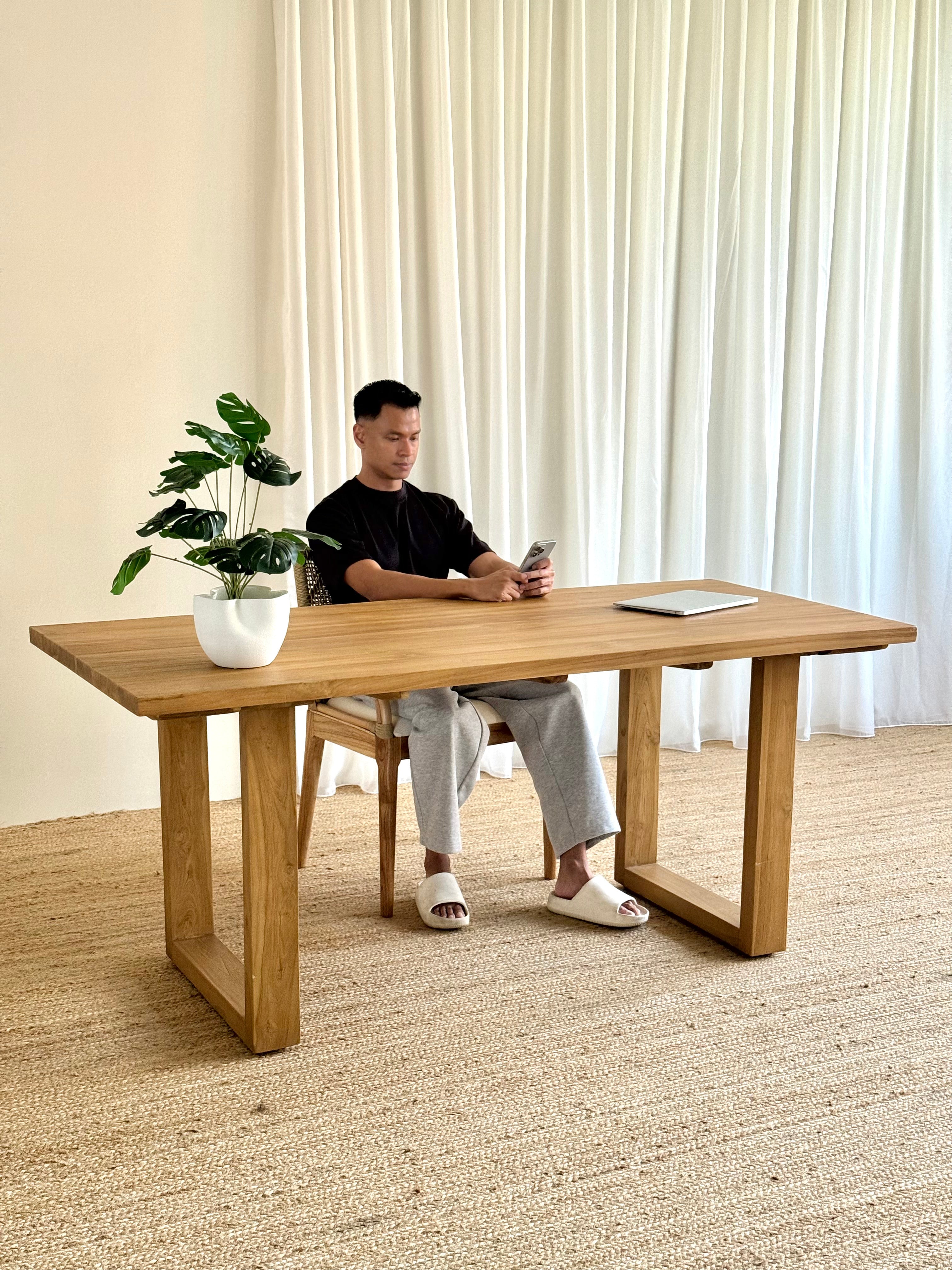 A man sits at a large light oak wooden writing desk in a bright room with cream curtains and a woven rug. The desk has a natural wood grain finish and blocky, geometric legs.