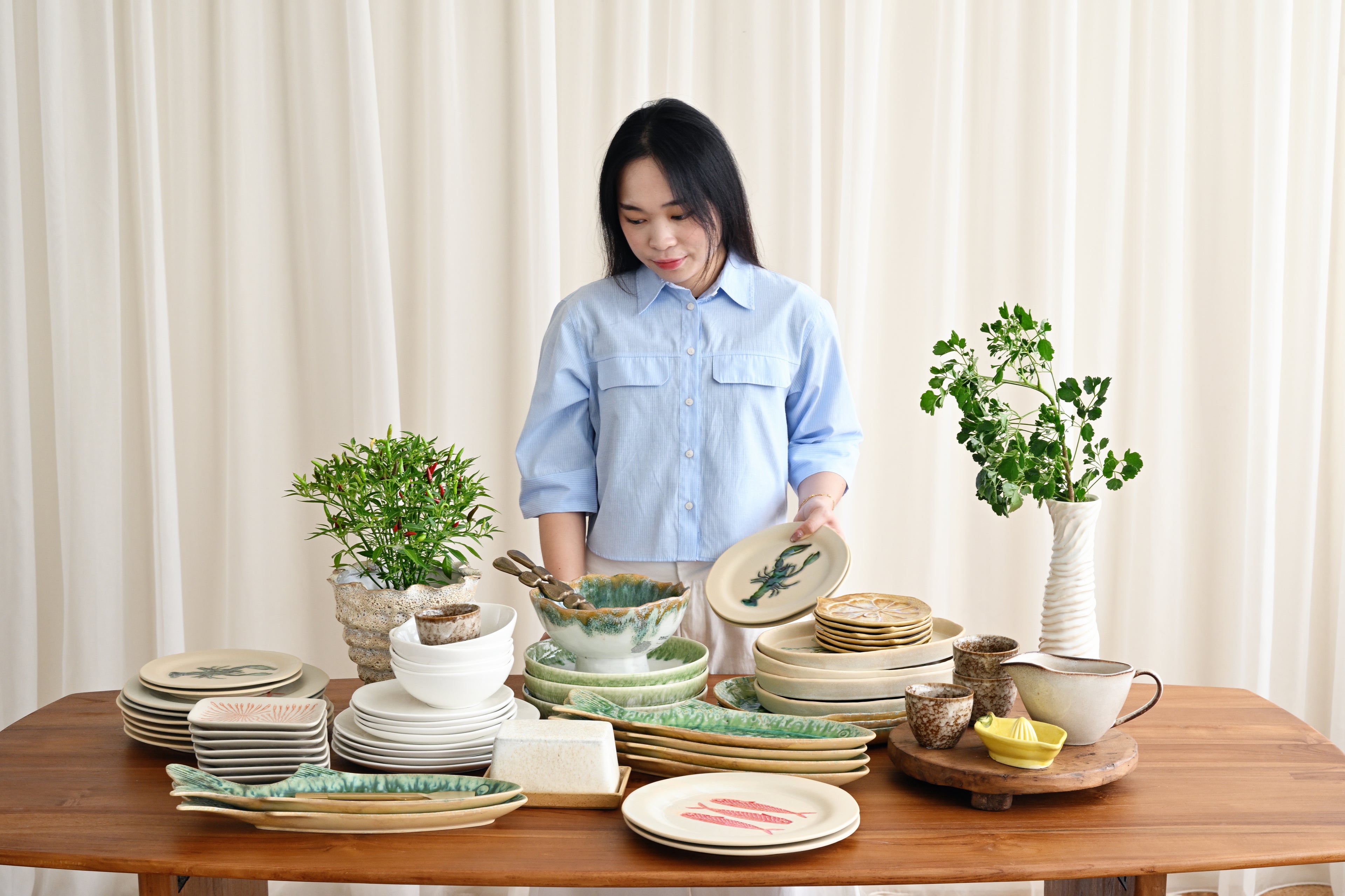 Woman arranging ceramic plates and bowls on a wooden table with plants in the background.