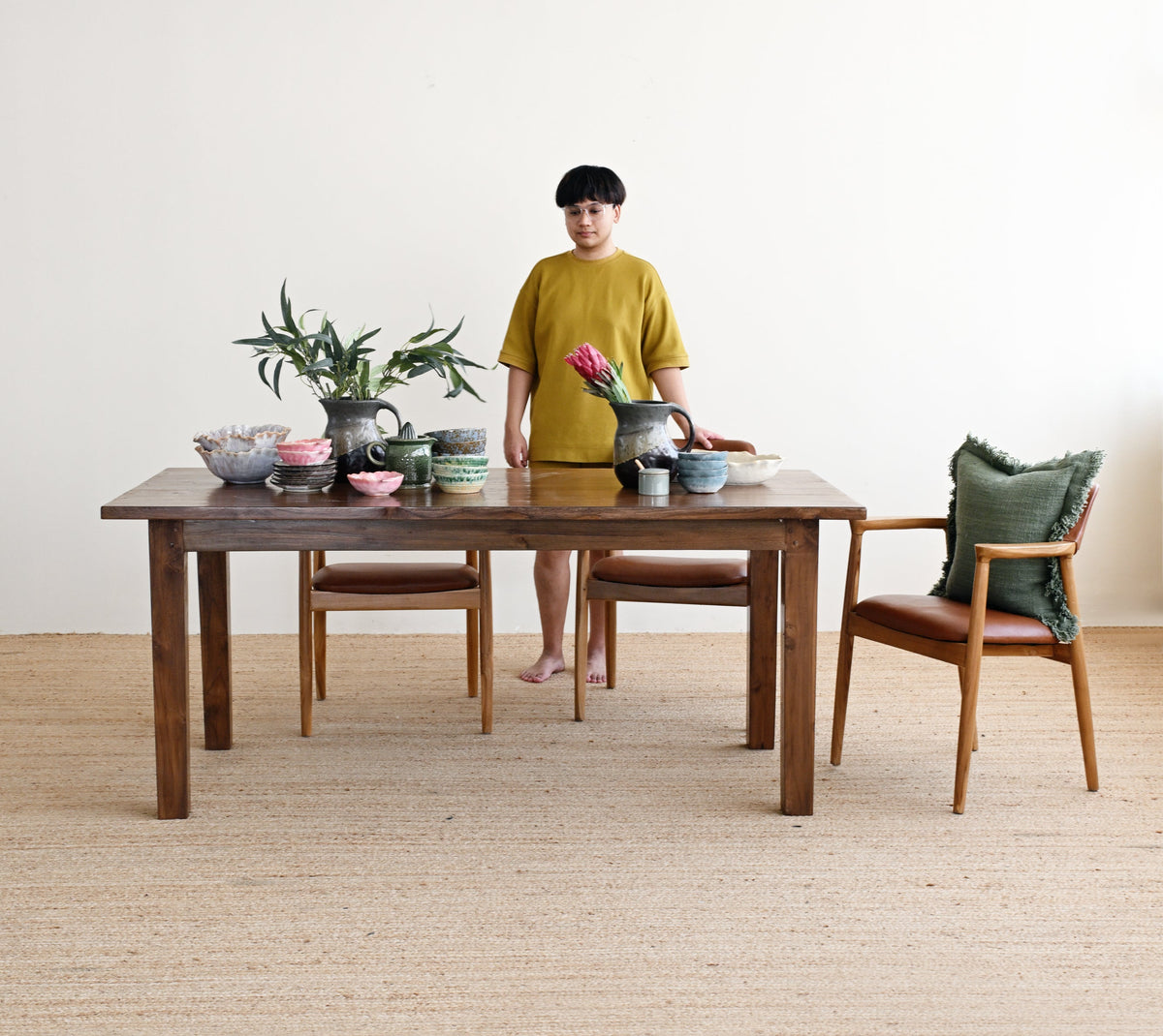 A person wearing a mustard yellow t-shirt and shorts stands behind a dark wood dining table set with bowls, a plant, and chairs in a minimalist room with a light beige wall and woven floor.