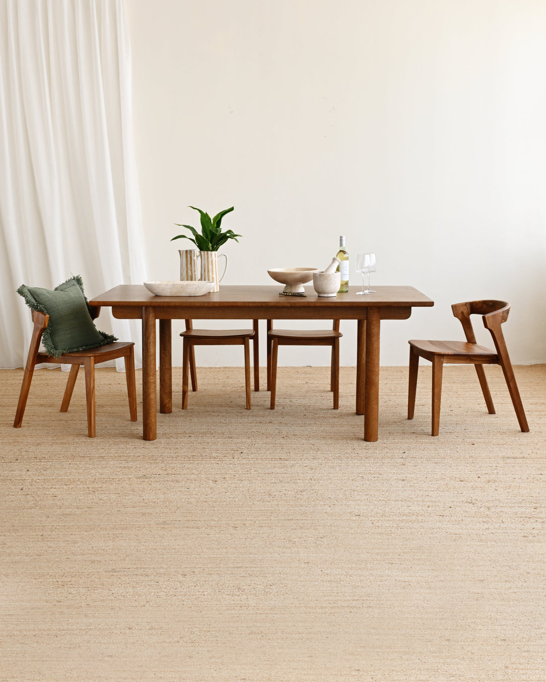 A mid-century modern wooden dining table set with four matching wooden chairs, styled with a small plant and ceramic dishes on a light beige woven rug against a neutral cream wall.