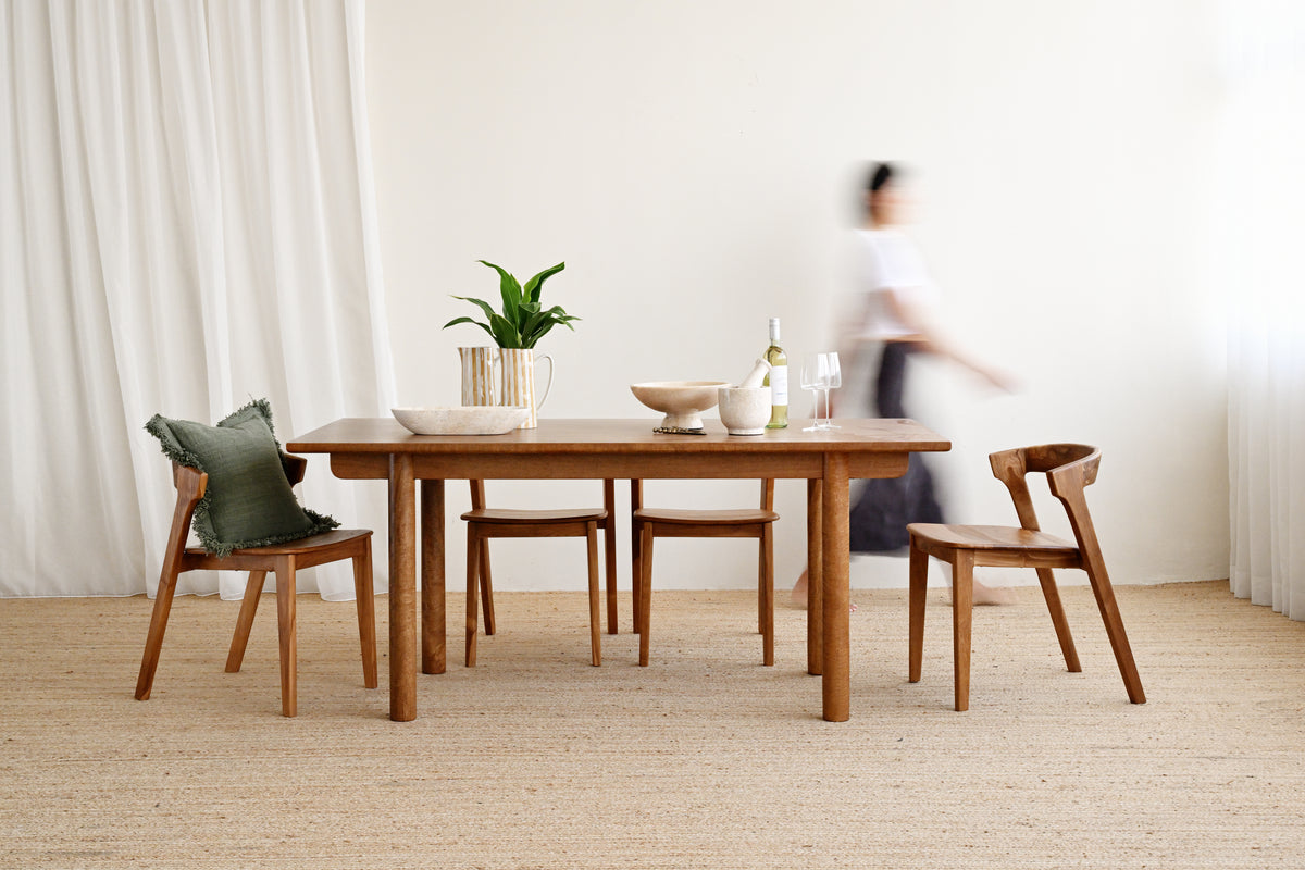 A wooden dining table with four matching wooden chairs in a bright, minimalist room with white curtains and a light beige rug. A green pillow rests on one chair, and a person is blurred in the background near the table.