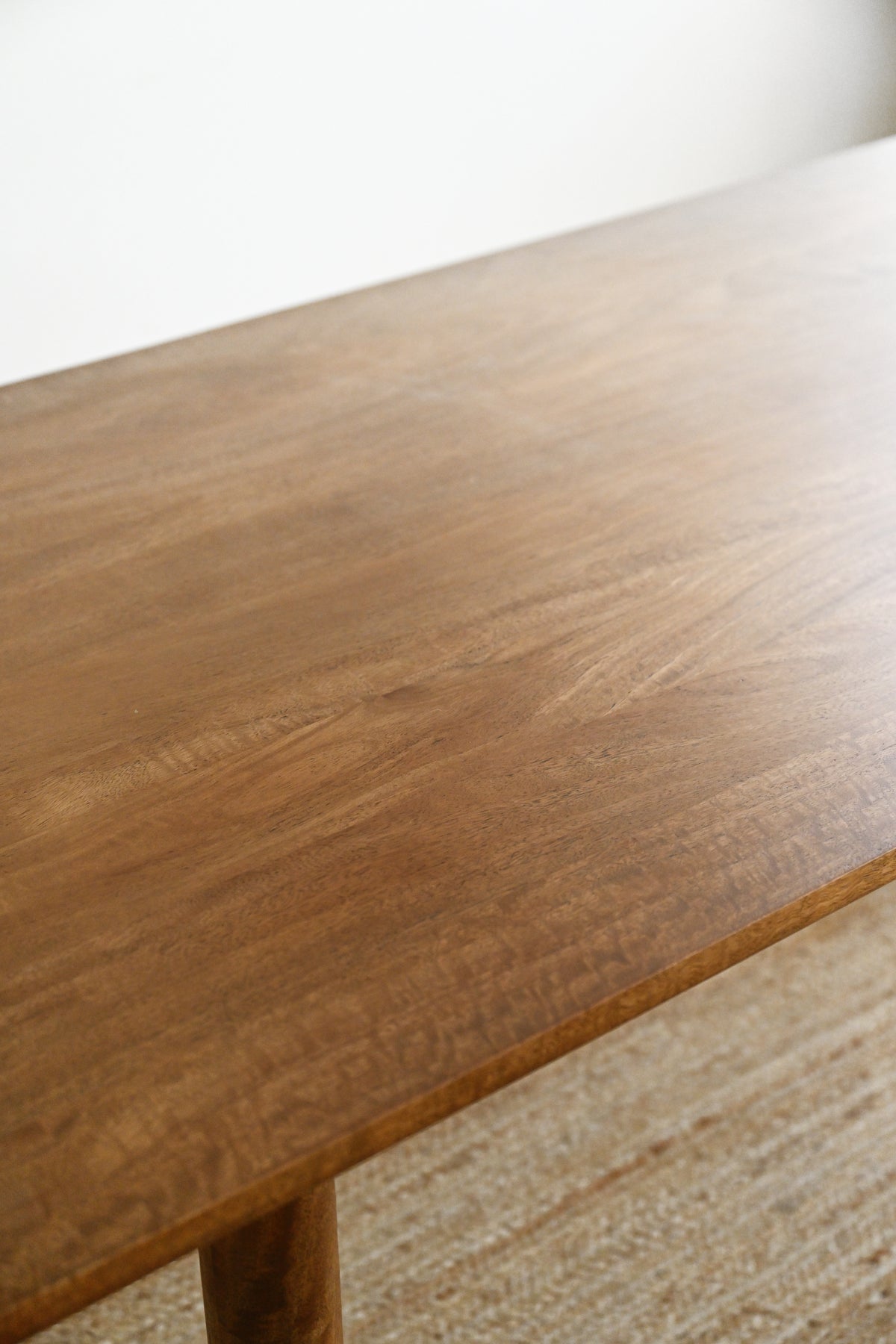 A close-up view of a warm medium-brown wooden table surface with visible wood grain, shown in a softly lit interior setting with a light neutral wall and woven rug.