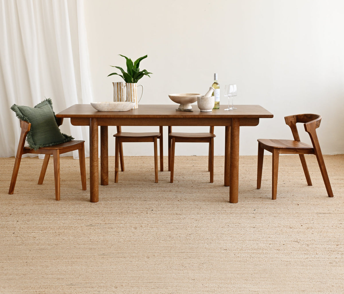 A mid-century modern wooden dining table set with four matching wooden chairs, styled with a small plant and ceramic dishes on a light beige woven rug against a neutral cream wall.