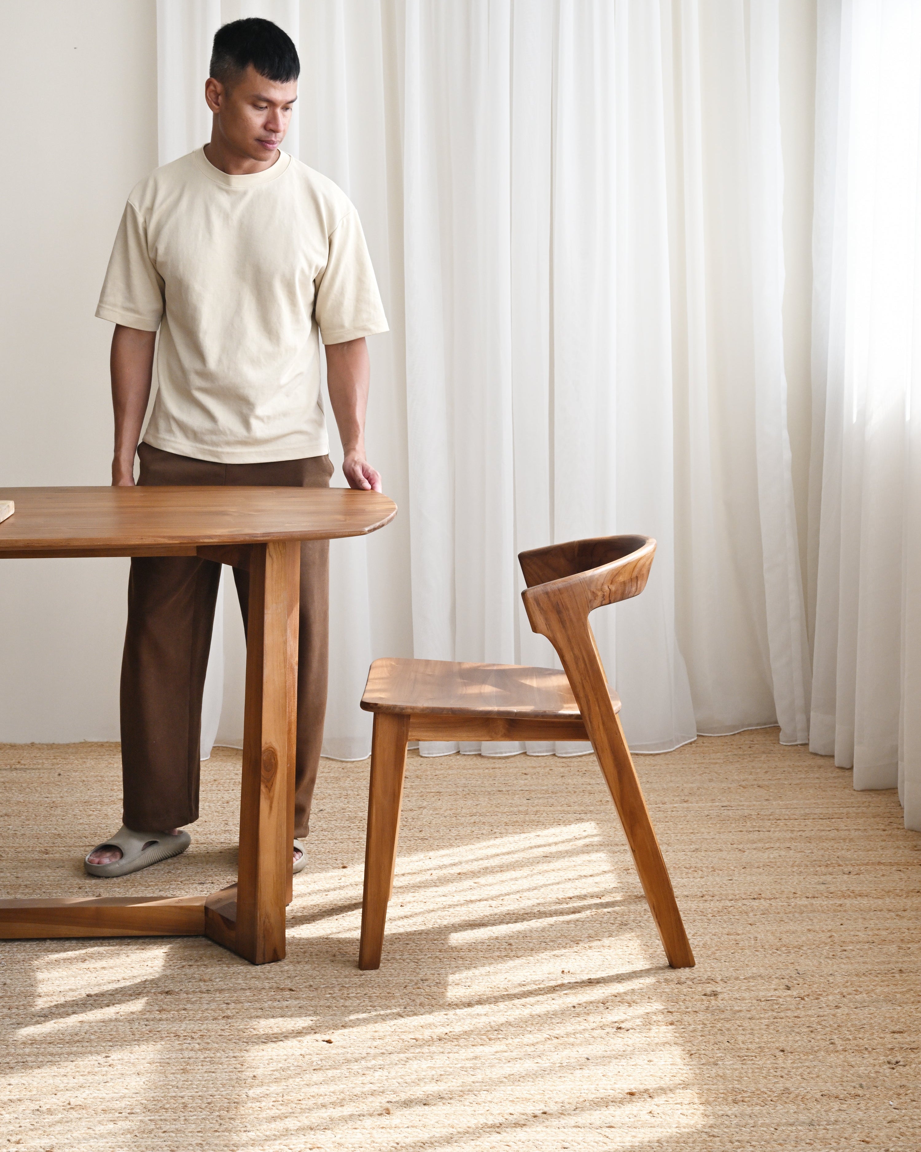 Man standing next to a wooden table and chair in a room with white curtains.