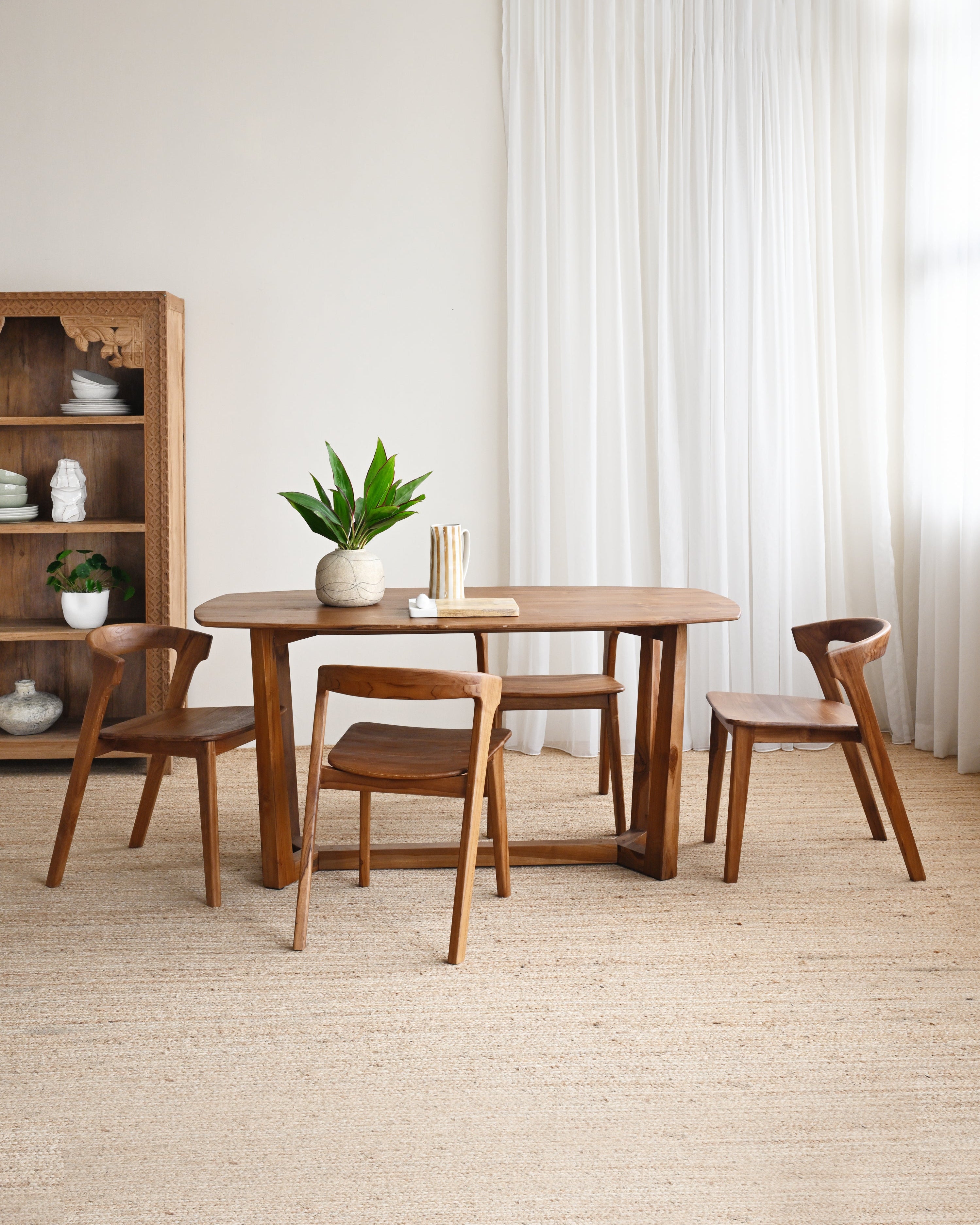 Dining area with wooden table and chairs in a room with white curtains and a bookshelf.