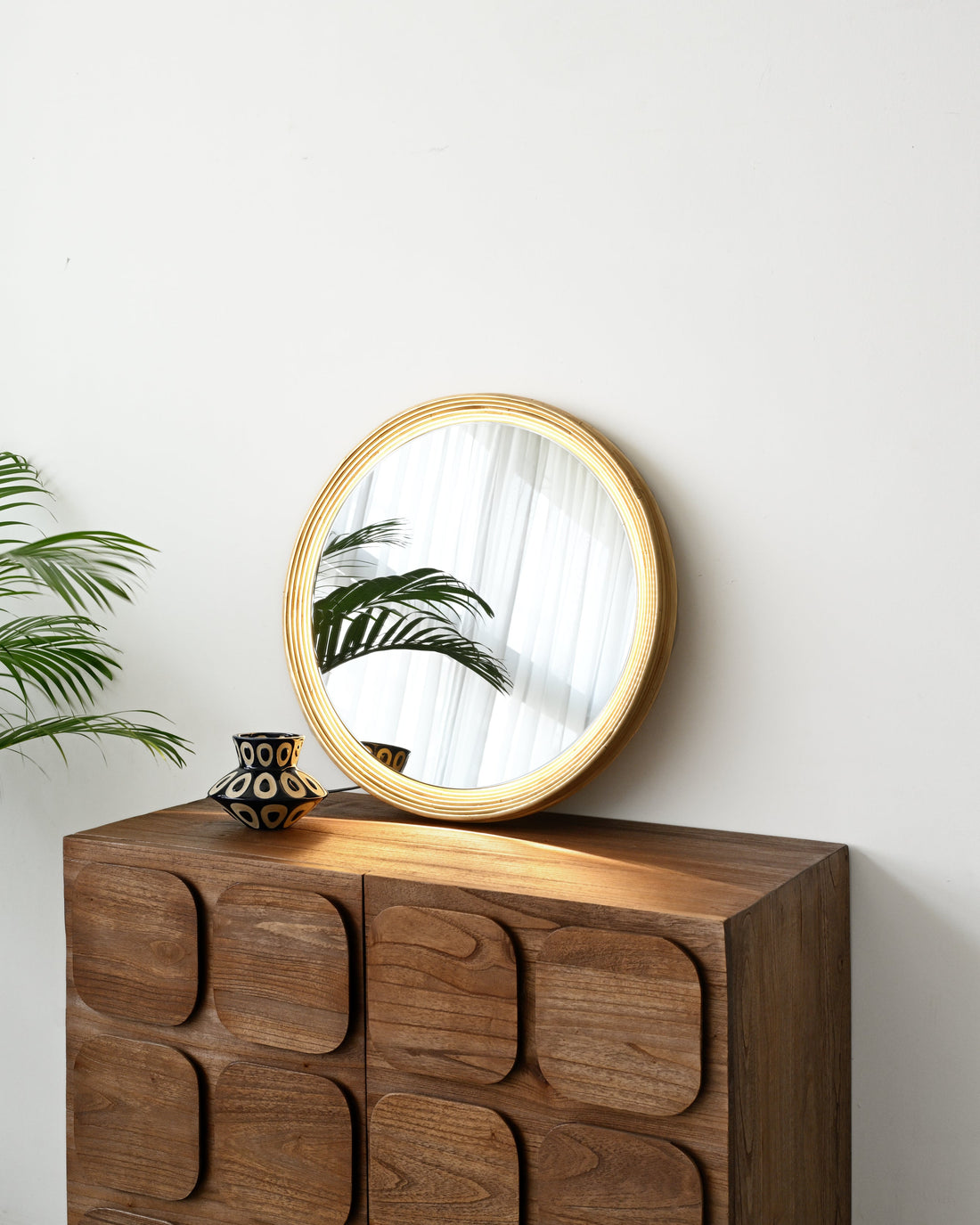 A round wooden-framed mirror with a gold-toned frame rests on top of a dark brown wooden cabinet with a unique raised geometric pattern, set against a plain white wall.
