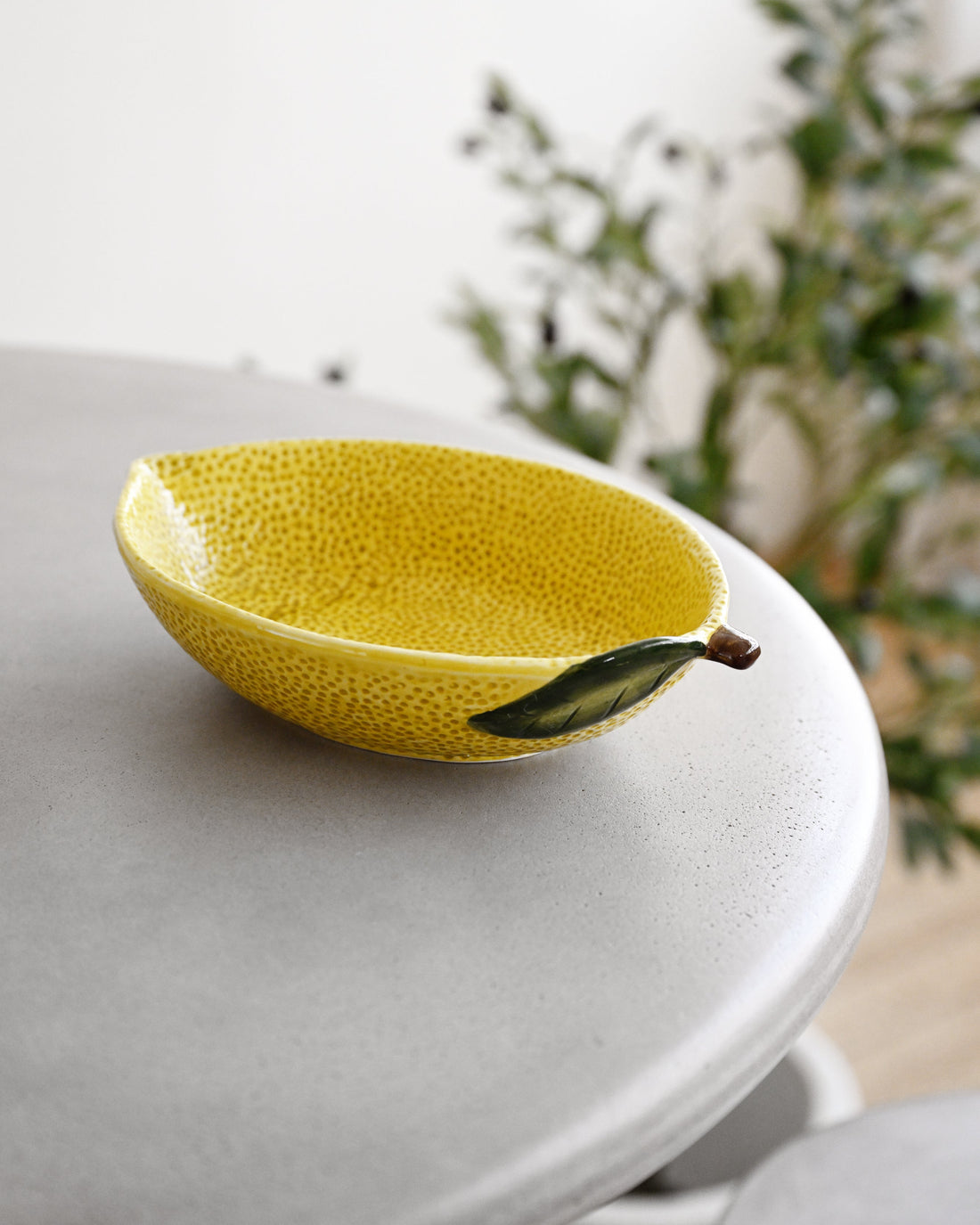 A yellow textured ceramic bowl designed to look like a lemon, featuring a green leaf detail on the side and a small stem, resting on a light gray table with a blurred indoor background.
